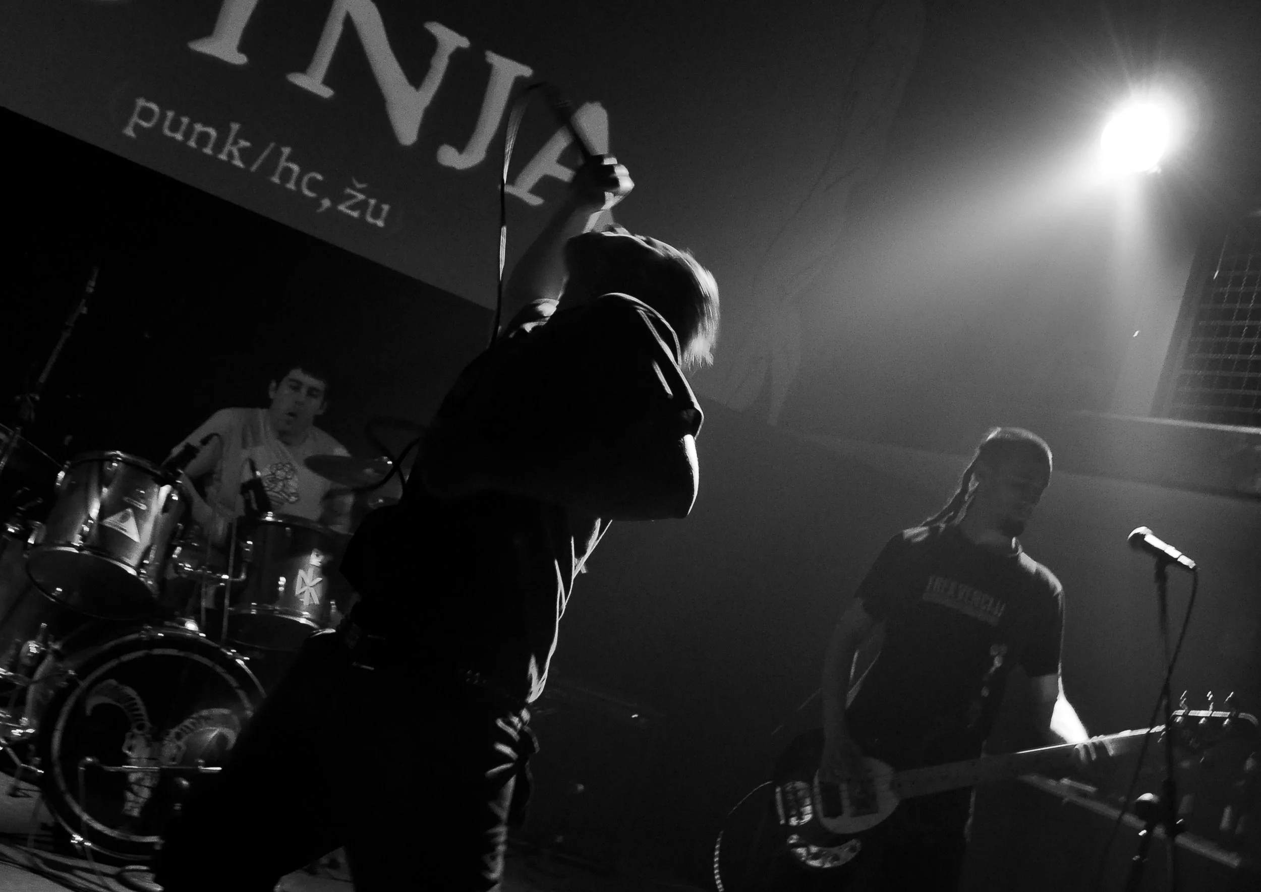 Black and white photo of a live music performance featuring a drummer, a vocalist, and a keyboard player on stage with a bright light shining from the right side.
