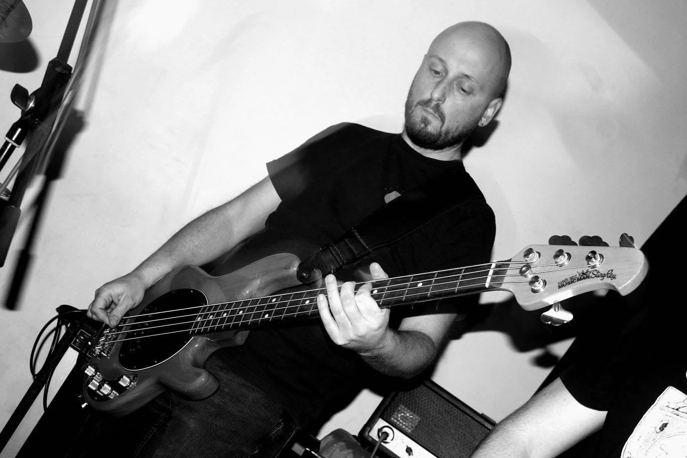 A man playing an electric bass guitar in a black shirt, in a room with musical equipment.