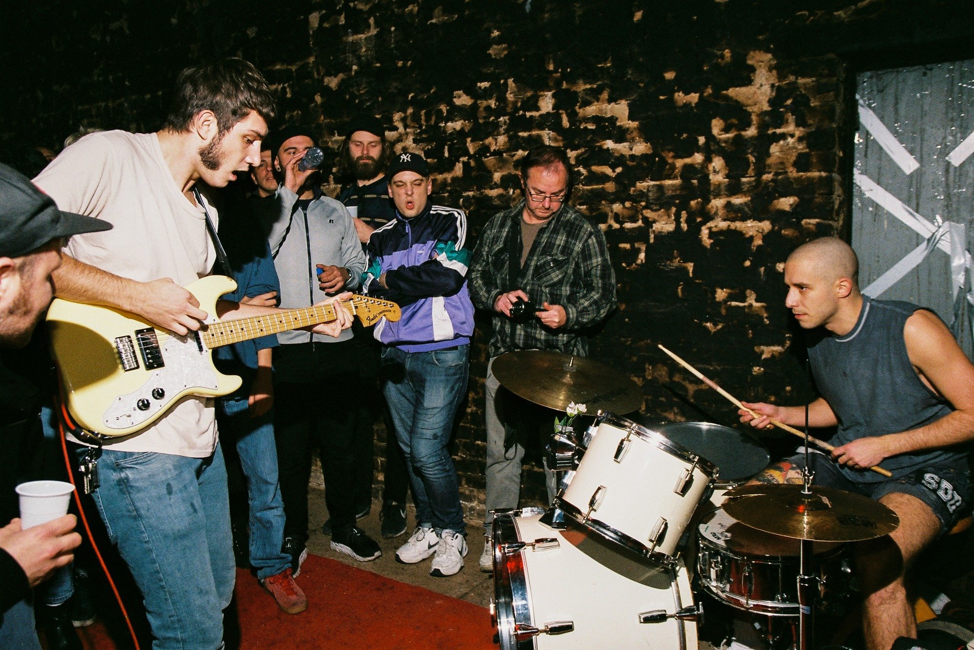 A group of people at a music jam session in a dimly lit room with exposed brick walls. One person is playing a yellow electric guitar, while another is playing the drums. Several others are watching, some with drinks in hand.