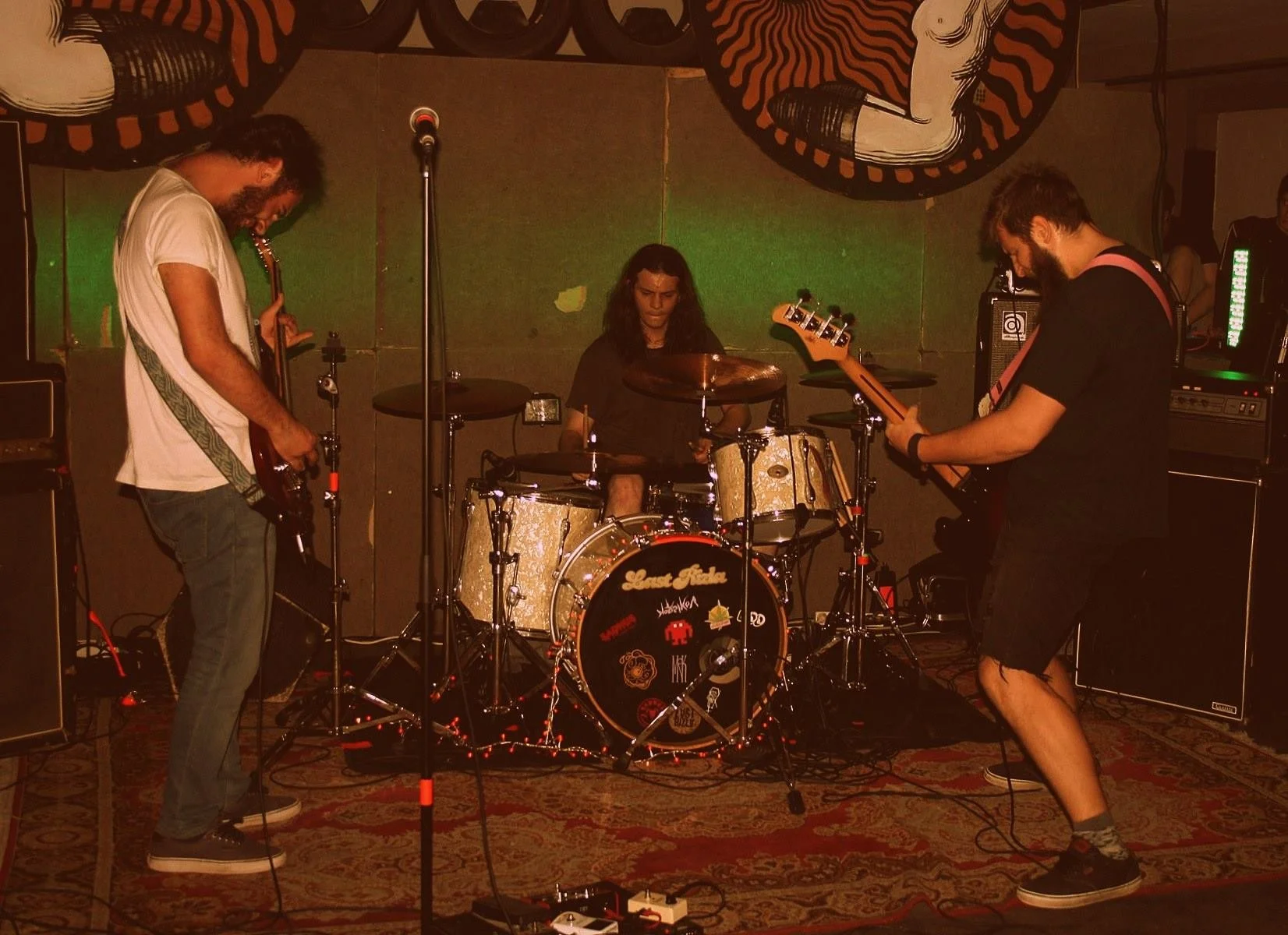 Three musicians performing on a stage with drums and guitars, in a dimly lit room with wall art and a decorative carpet.