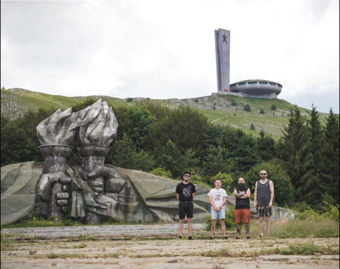 Four men standing in front of a monument with a sculpted image of hands holding a torch, and a large building with a star on it on a hill in the background.