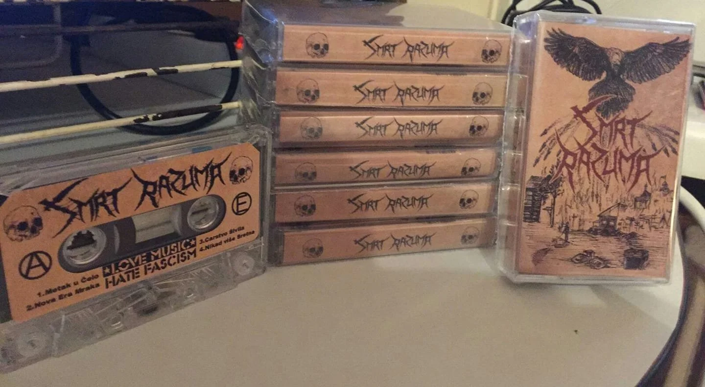 A cassette tape titled 'Smrt Raz' with pink cover art featuring a black eagle holding a cross and a town scene, stacked with eight other identical small cassette tapes, and another cassette case with matching cover art leaning on a plastic container.