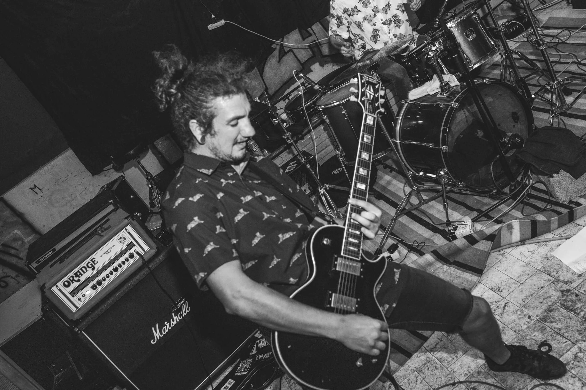 A musician playing an electric guitar in front of an Orange guitar amplifier, with drums behind him in a music studio or practice space.