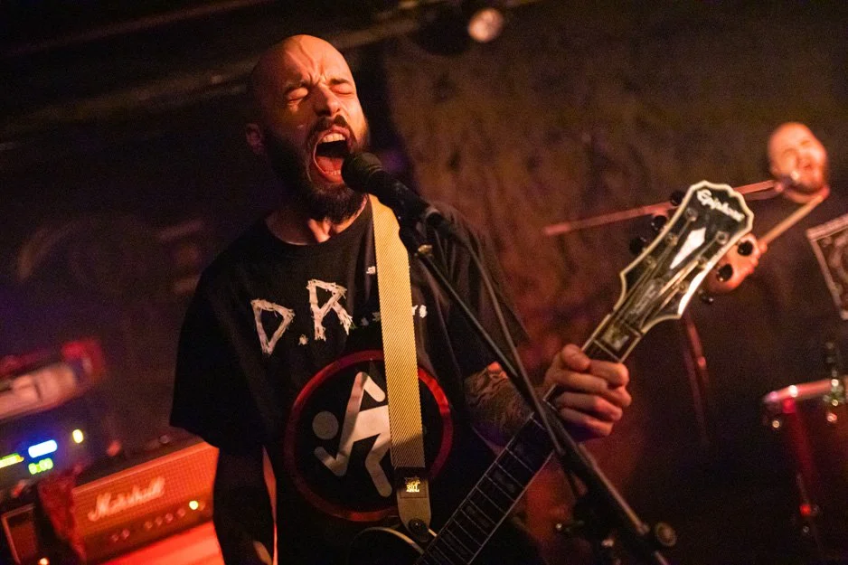 A man passionately singing into a microphone while playing an electric guitar on stage in a dimly lit music venue, with another musician in the background playing the drums.