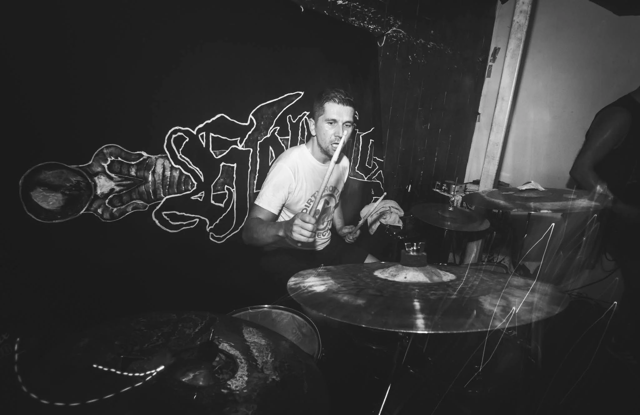 A man playing drums in a dimly lit room with a dark wall behind him. There is neon art on the wall and a drum set in front of him.