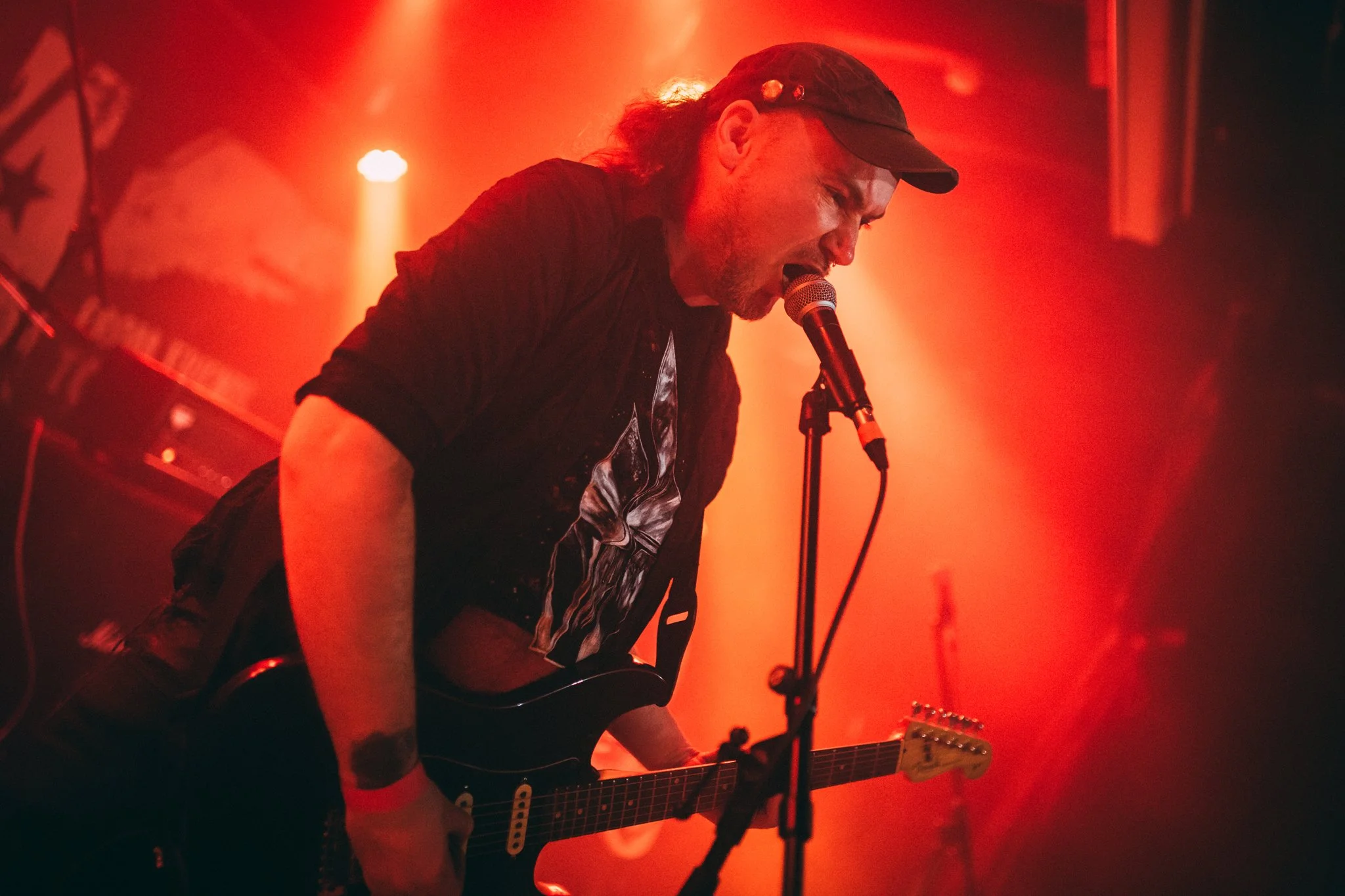 Musician singing into a microphone and playing an electric guitar on stage with red lighting.