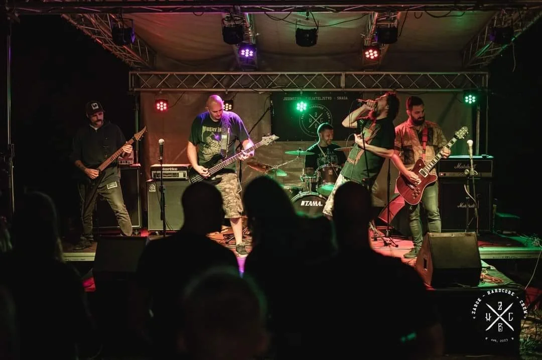 A band performs live on stage with four musicians playing electric guitars and bass, a drummer, and a vocalist singing into a microphone, with colorful stage lighting and an audience watching.