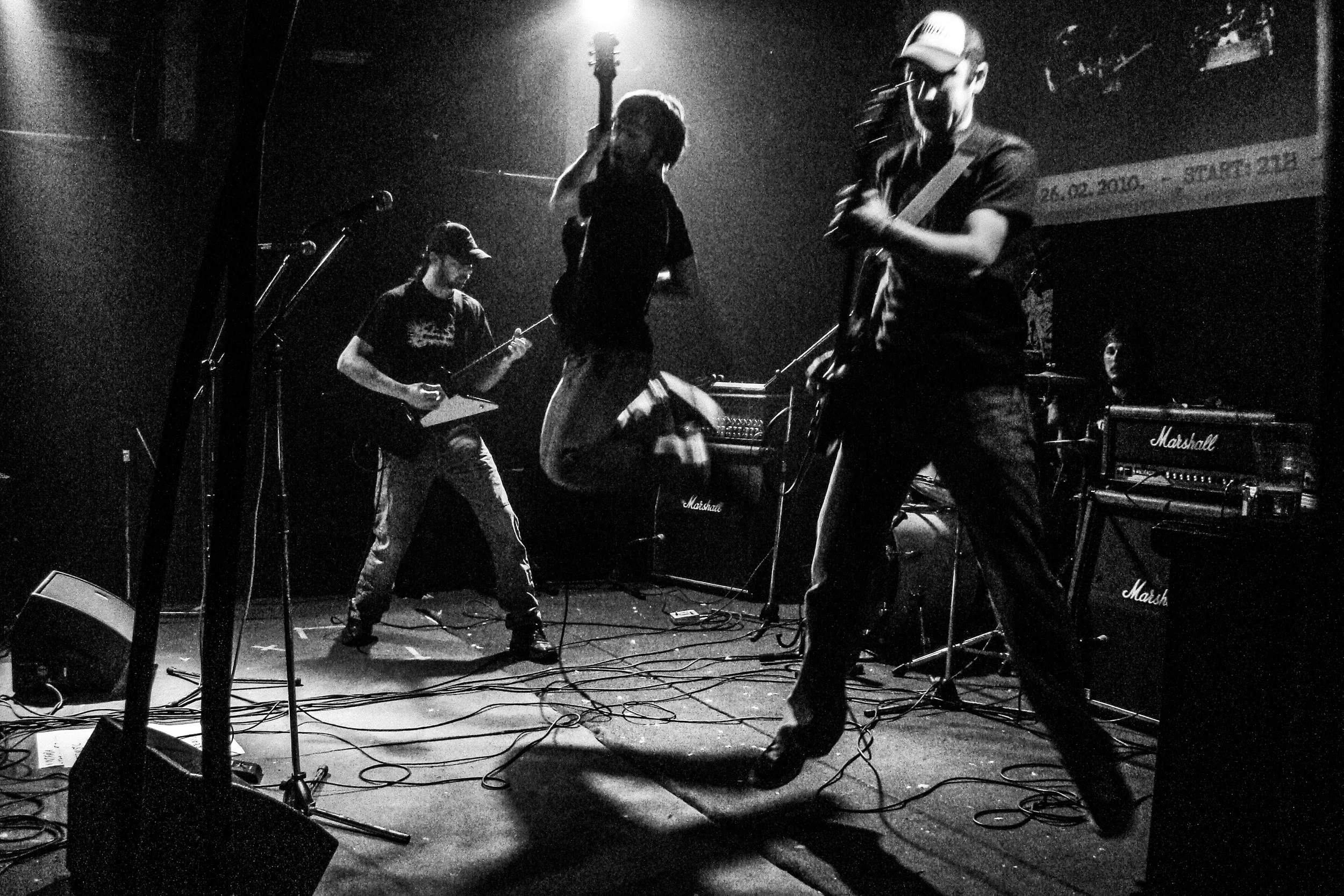 A black and white photo of a band performing on stage, including a vocalist mid-air, a guitarist, a bassist, and a drummer in the background, with various musical equipment like microphones, amplifiers, and cables on the stage.