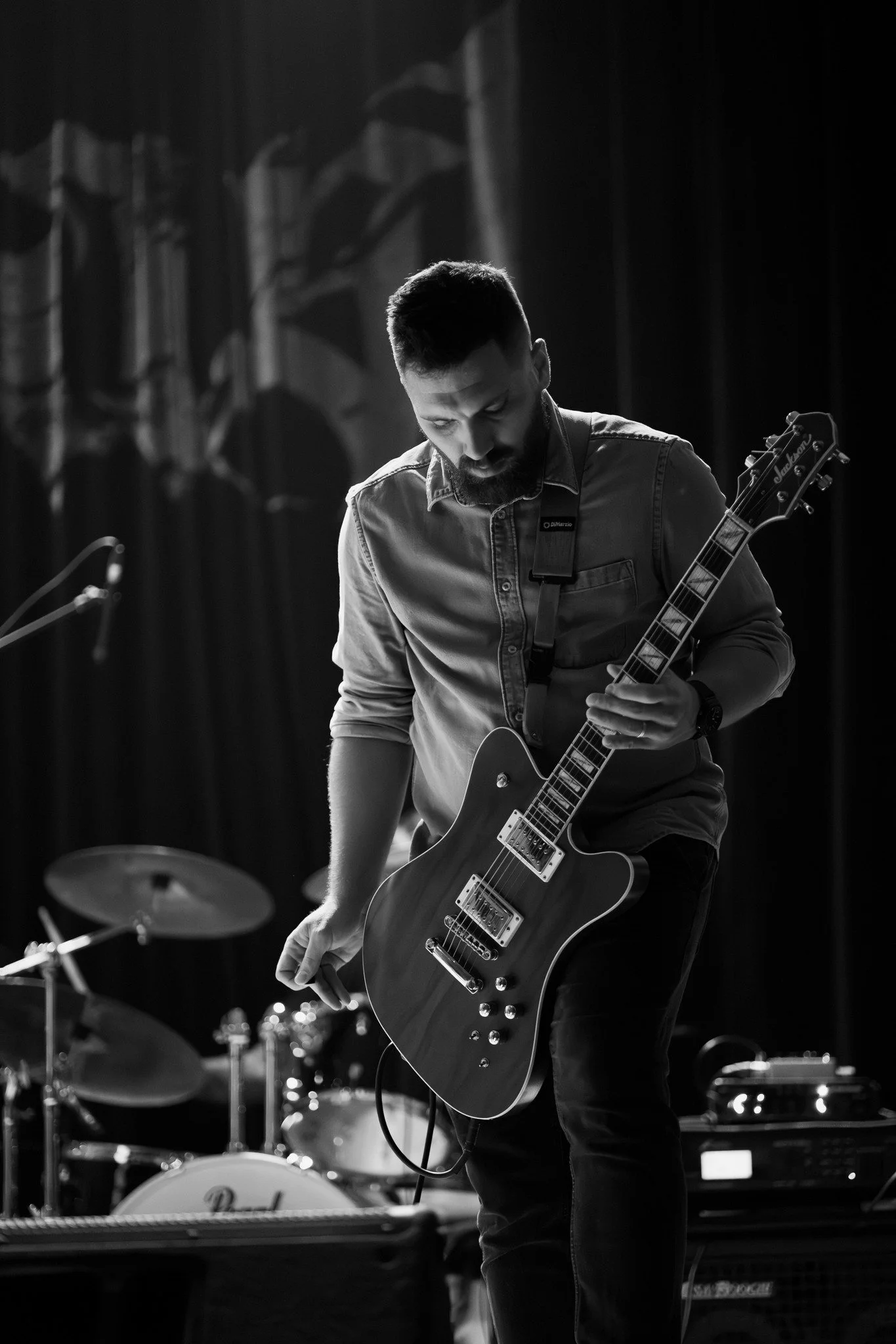 Black and white photo of a man with short hair and a beard playing an electric guitar on stage, with drums in the background.