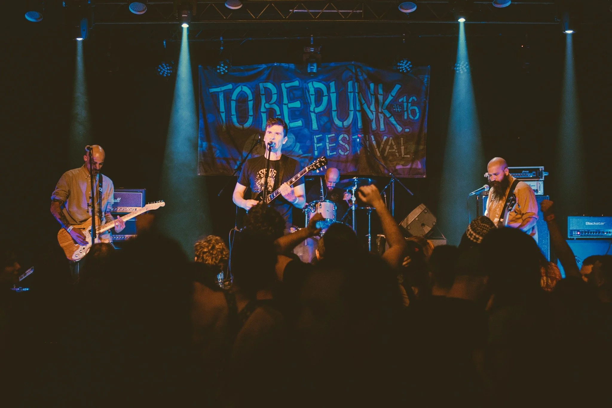 A band performing live on stage at To be punk Festival. The lead singer is playing an electric guitar, with other band members playing guitars and drums. The stage is lit with blue lights and a large banner with the event's name hangs at the back.