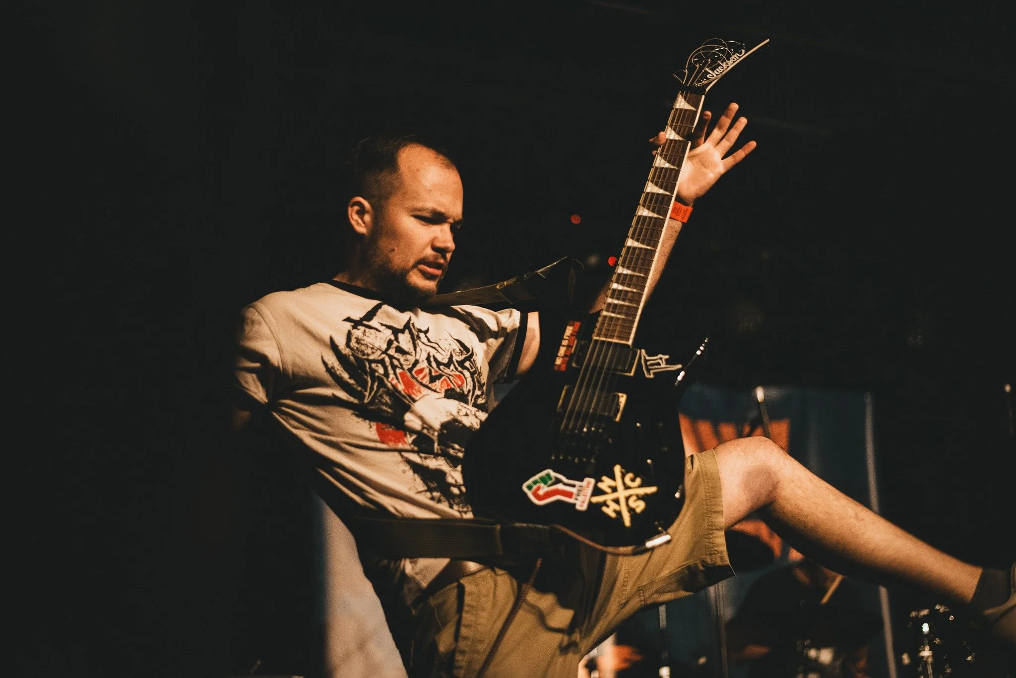 A man sitting in a dark room playing an electric guitar, wearing a beige and black t-shirt and khaki shorts, with his eyes closed and showing a focused expression.
