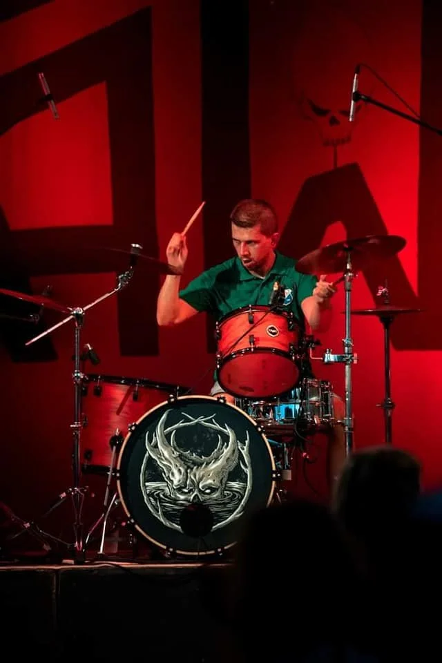 A man playing a red drum set on stage with a black and white deer skull graphic on the bass drum, under red stage lighting.