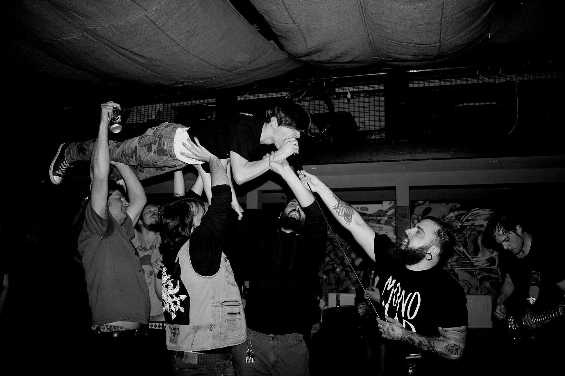 A group of people at a concert or live music event, with a man with a beard and tattoos holding a microphone, another person elevated above others, possibly during a crowd surfing moment, and others watching and enjoying the performance.