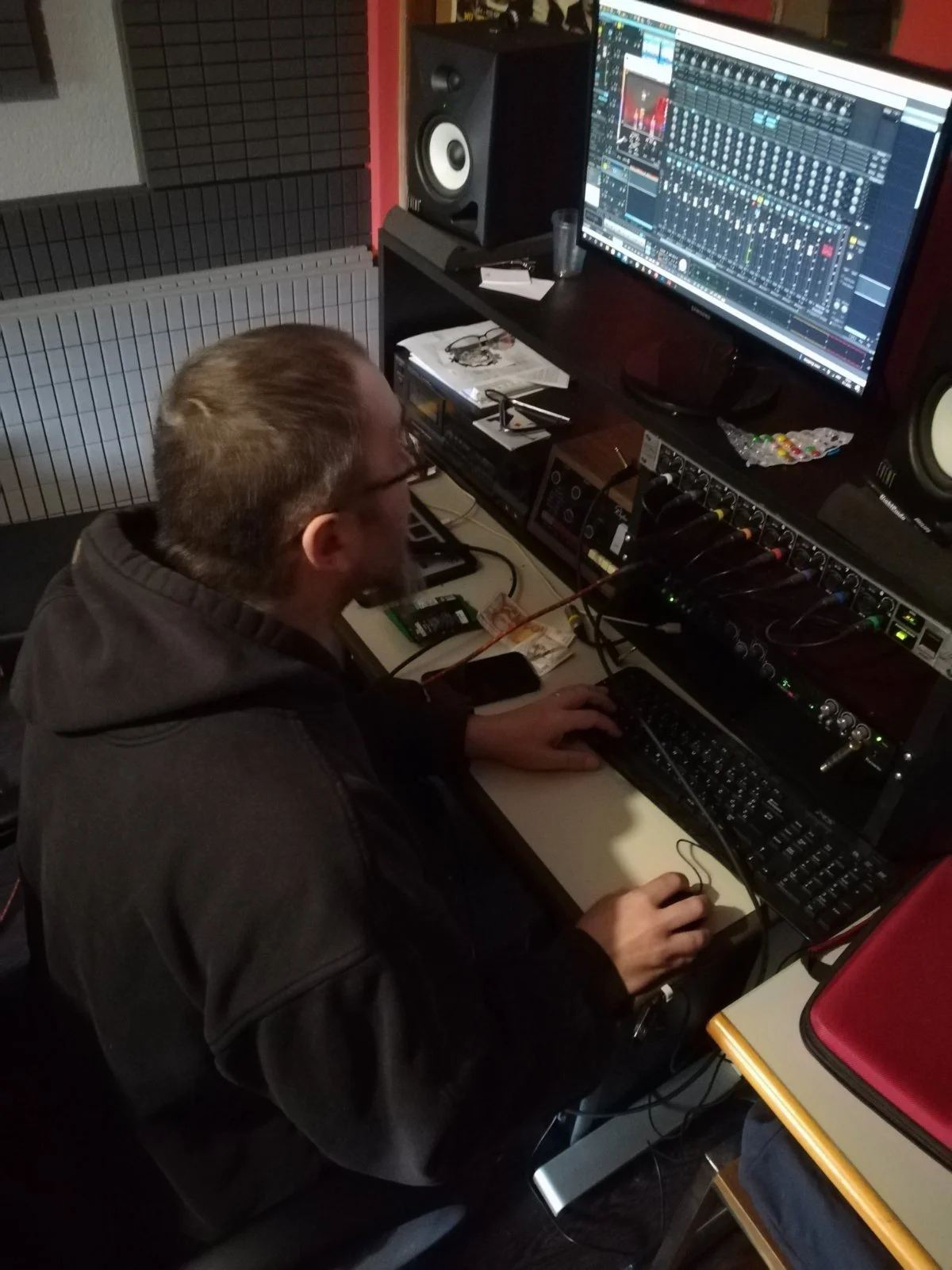 A man with glasses and a beard working at a music production station with computer screens displaying audio editing software, studio monitors, and audio equipment.
