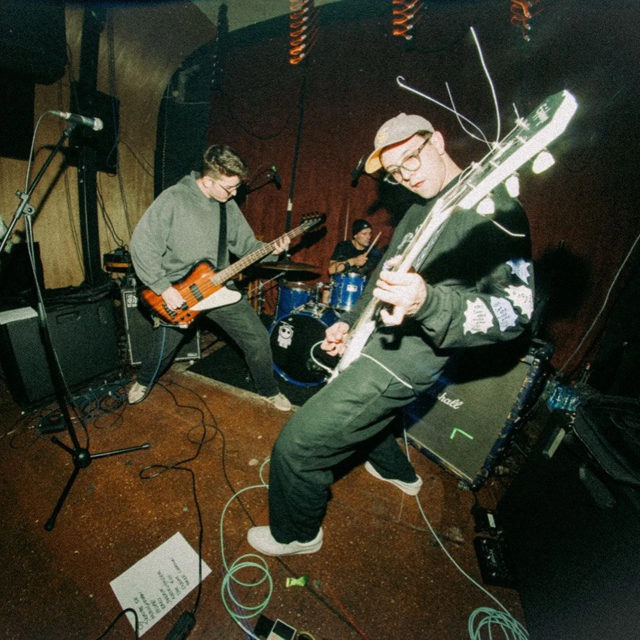 A band performing on stage with three members: a guitarist with glasses and a hoodie, a bassist with a bass guitar, and a drummer with drumsticks in hand. Various equipment and cables are visible on the stage.