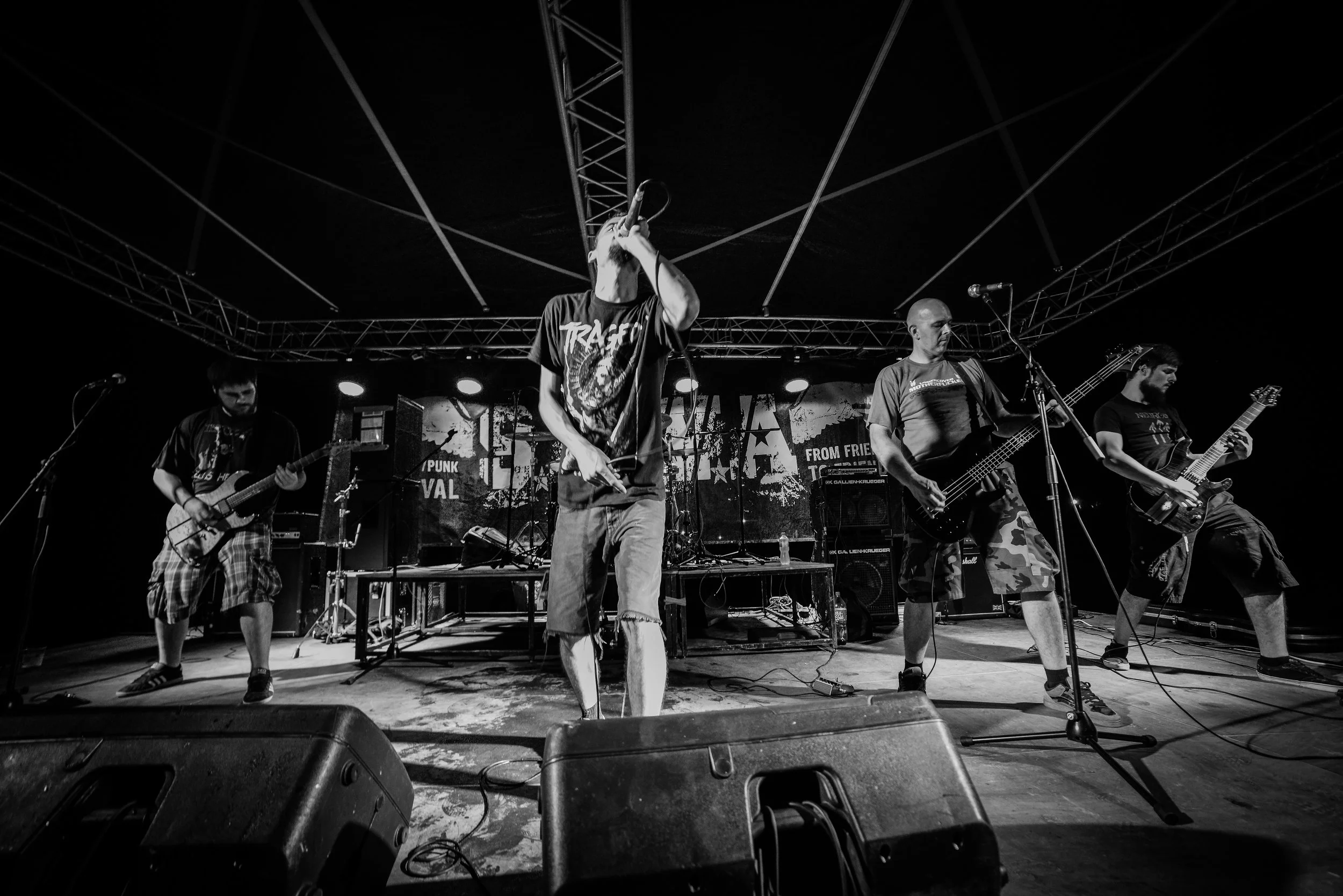 A black and white photo of a band performing on stage, featuring four members playing guitars and singing. The stage has equipment, microphones, and a backdrop with text. Band name: Proslov