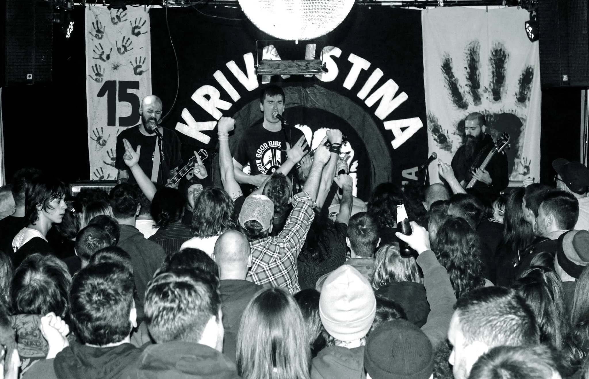 A black and white photo of a live music performance of a band called 'Kriva Istina. The band is playing on stage with a crowd of people in front. The stage has a backdrop with the venue's name and handprints.