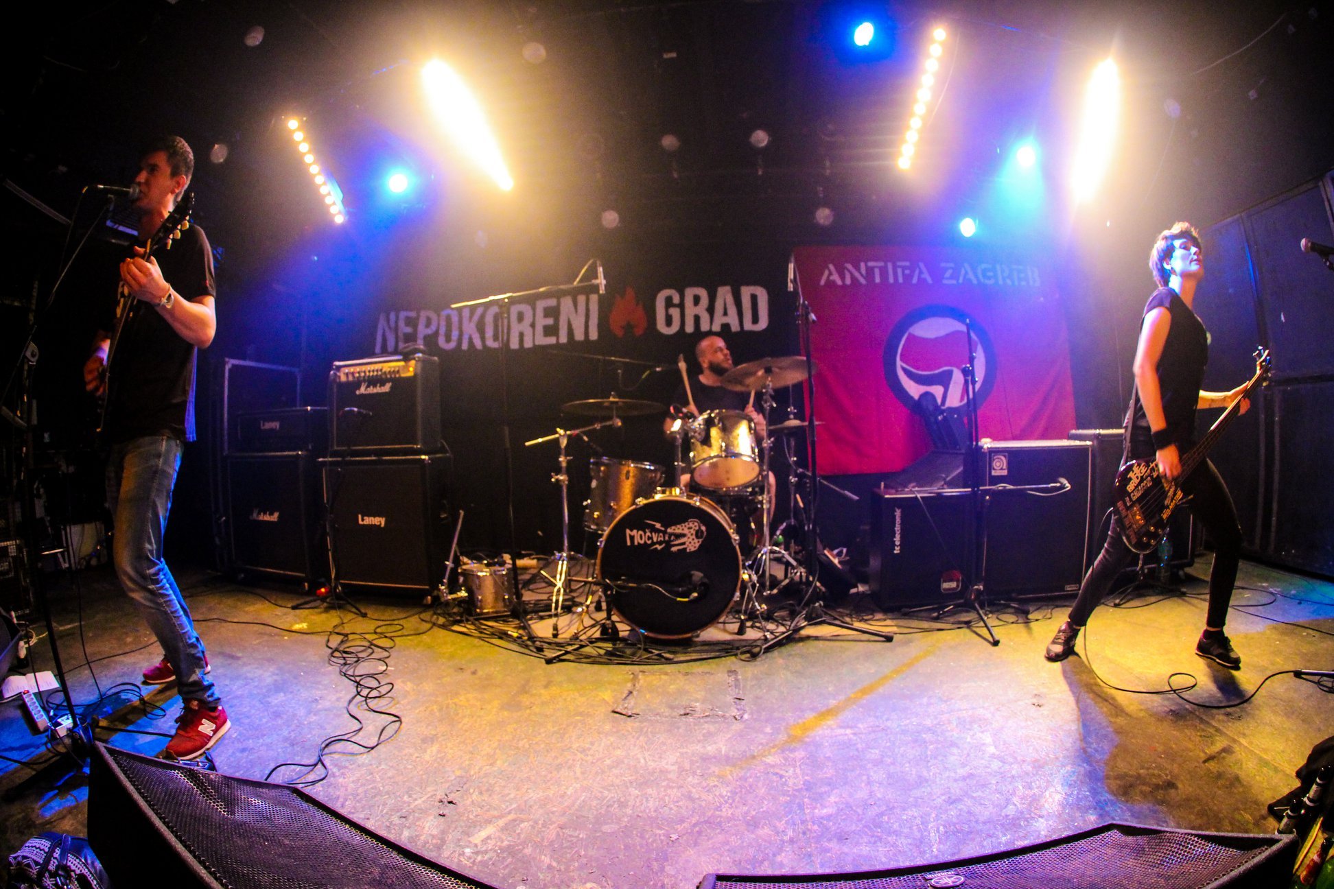 Band performing on stage with instruments including guitar, bass, and drums, in a venue decorated with banners featuring Balkan themes that read "Nepokorni Grad" and "Antifa Zagreb," colored stage lighting and sound equipment visible.
