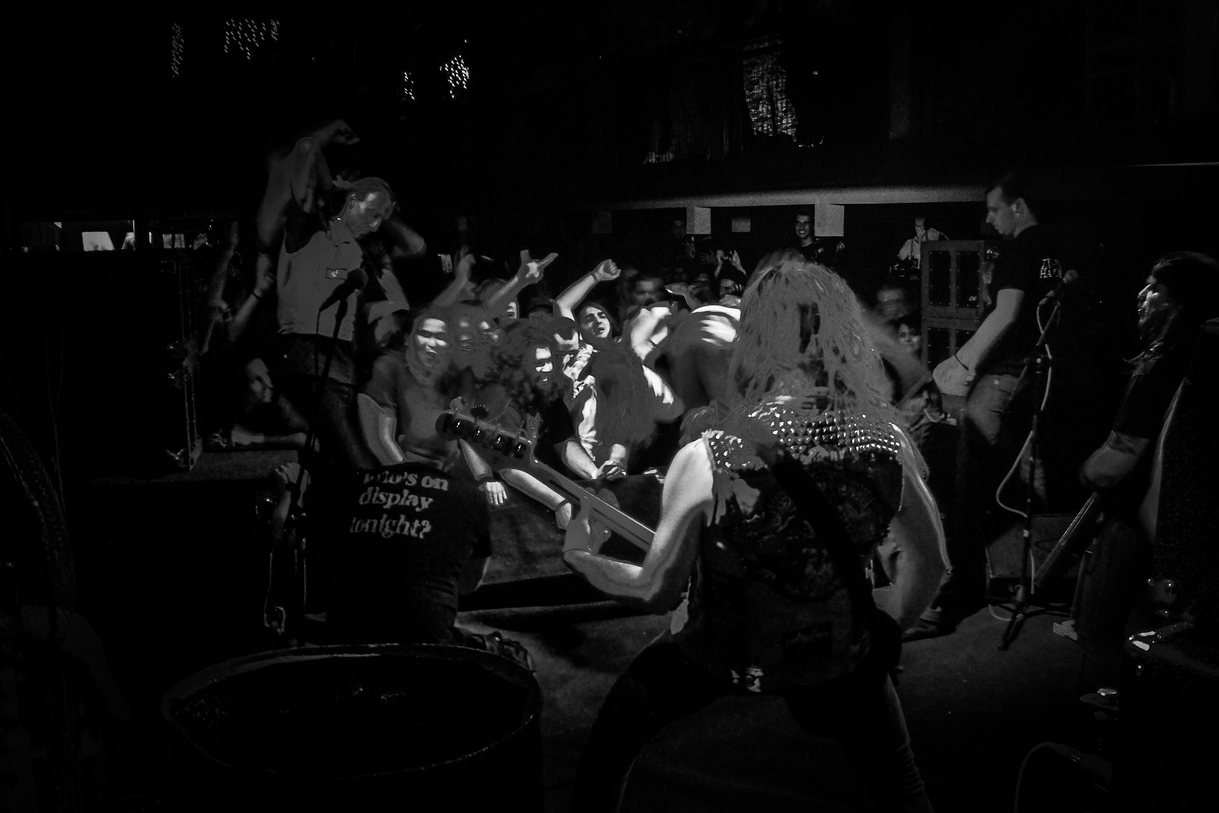 A crowd of people at a concert or party, with some raising their hands and enjoying live music, with performers on stage and a DJ in the background.