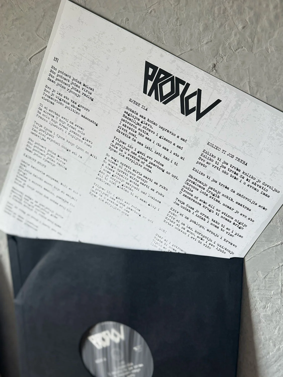 A partially opened black envelope containing a printed sheet of paper with a stylized black logo or title, and text written in Serbian or Croatian, placed on a textured gray surface.