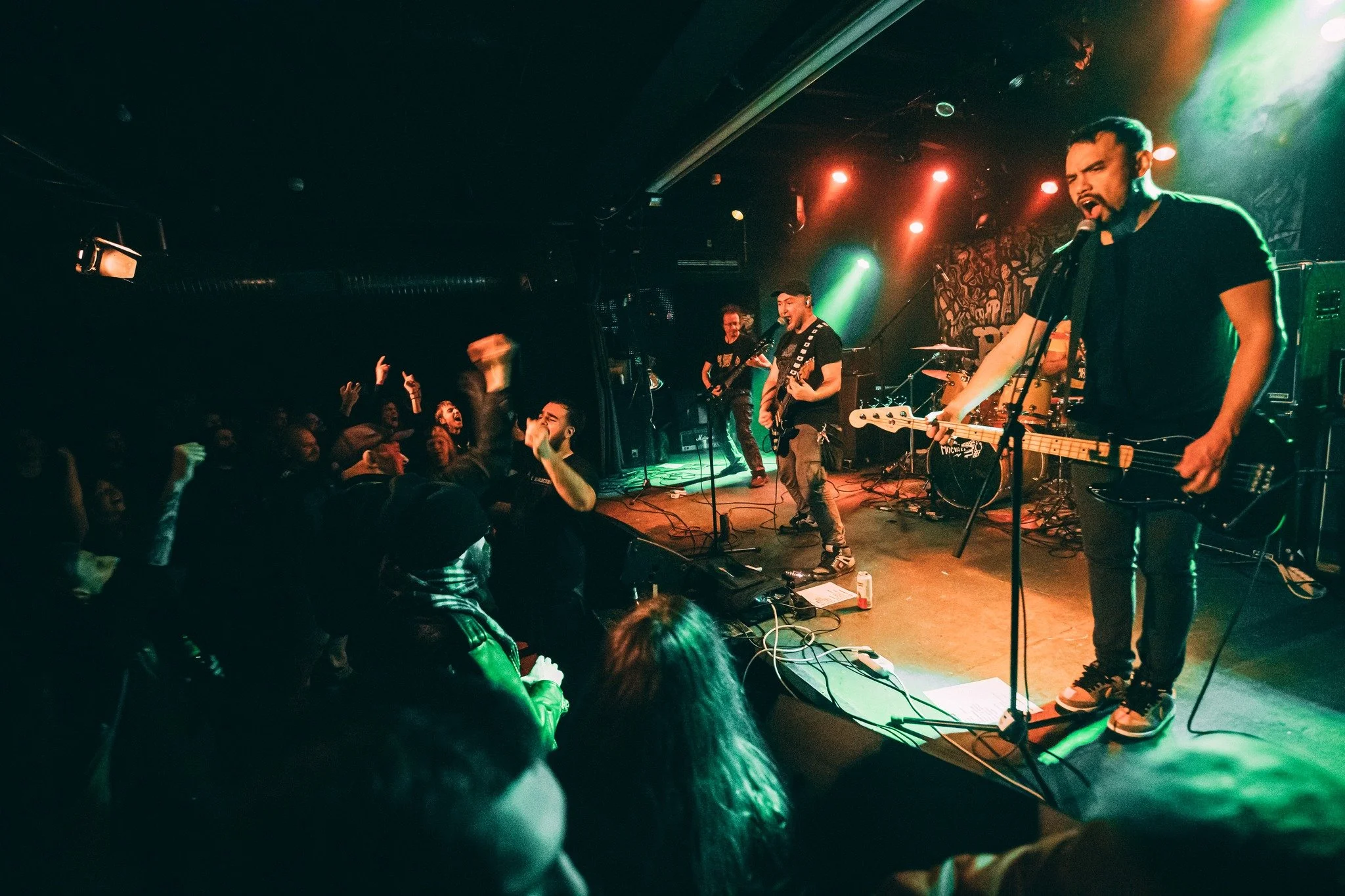 A band performing live on stage in a dark venue, with the crowd cheering in front and colorful stage lights illuminating the scene.