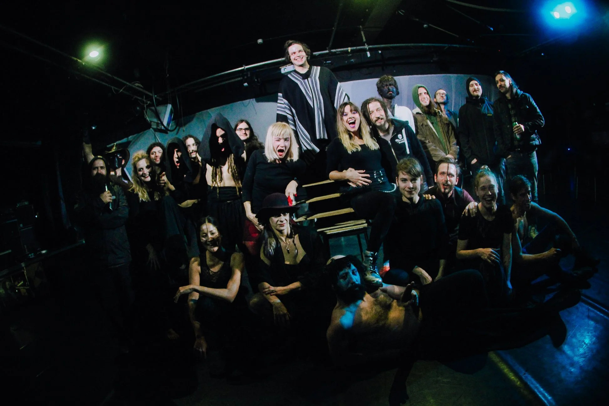 Group of people on stage at a concert or performance venue, smiling and posing for a photo. Stage lights and equipment are visible in the background.
