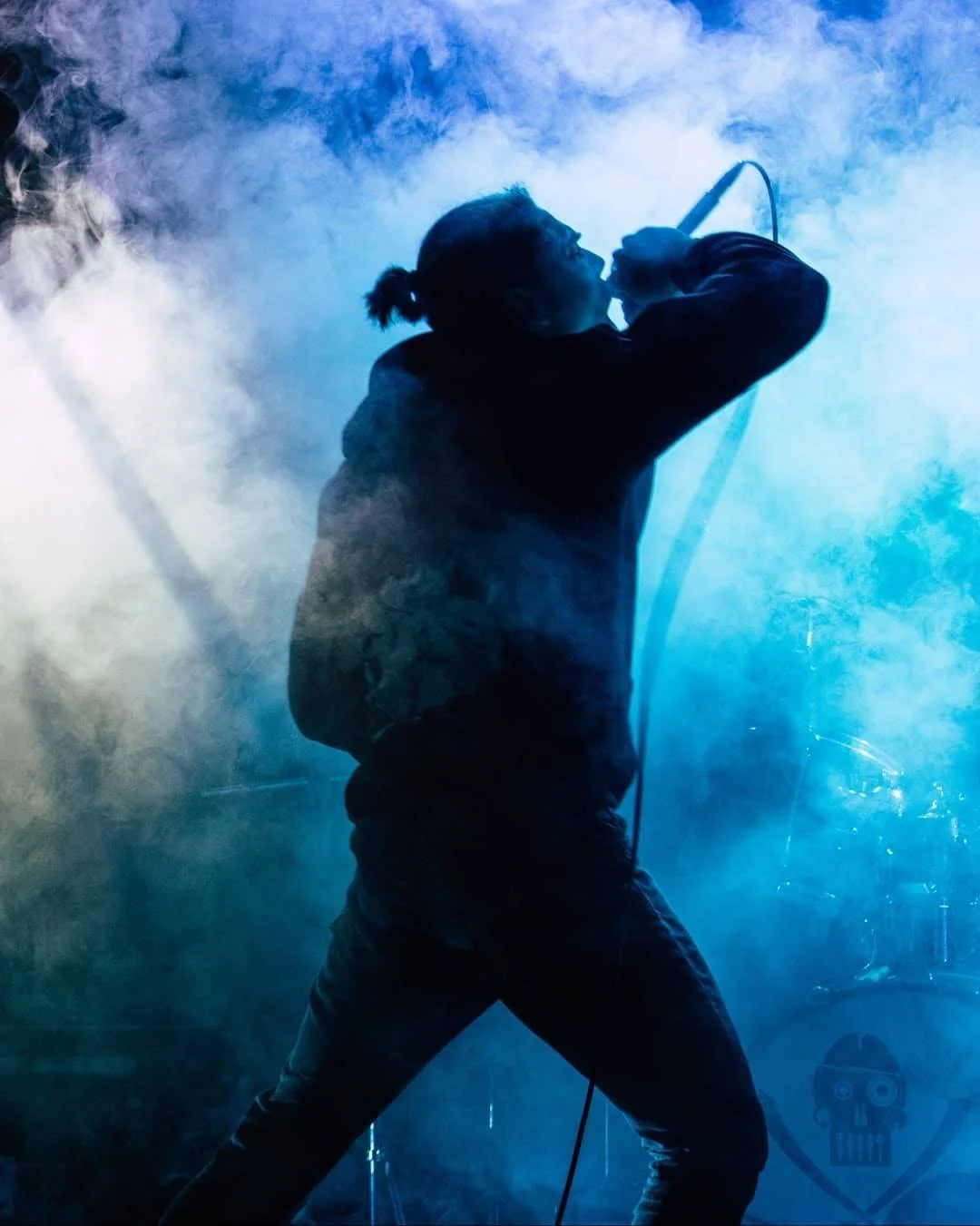 A silhouette of a singer performing on stage, holding a microphone, surrounded by blue and white stage smoke.