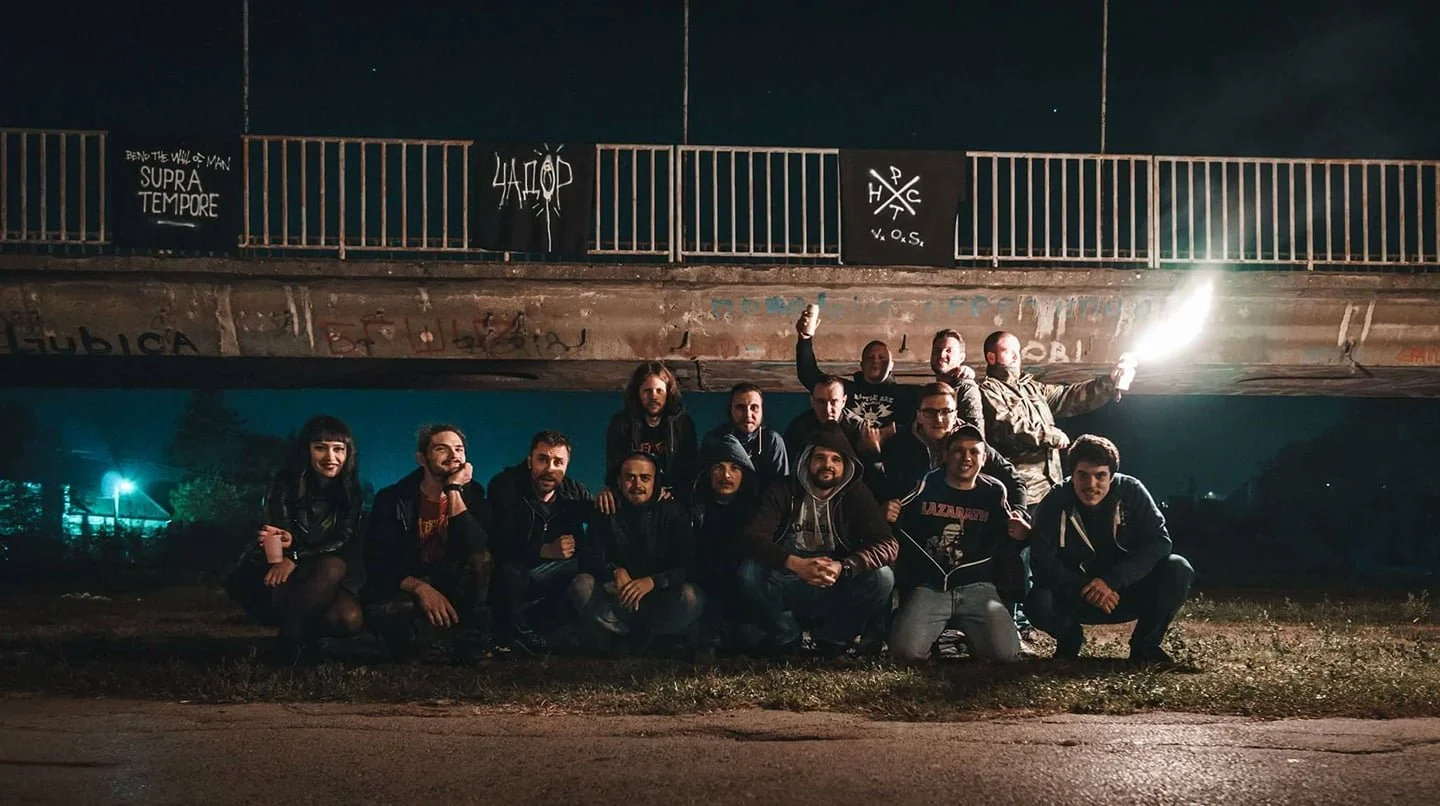 Group of about twelve young adults posing outside at night, some holding flares, beneath a bridge with graffiti and banners. Some are kneeling, some standing, with a dark sky in the background.
