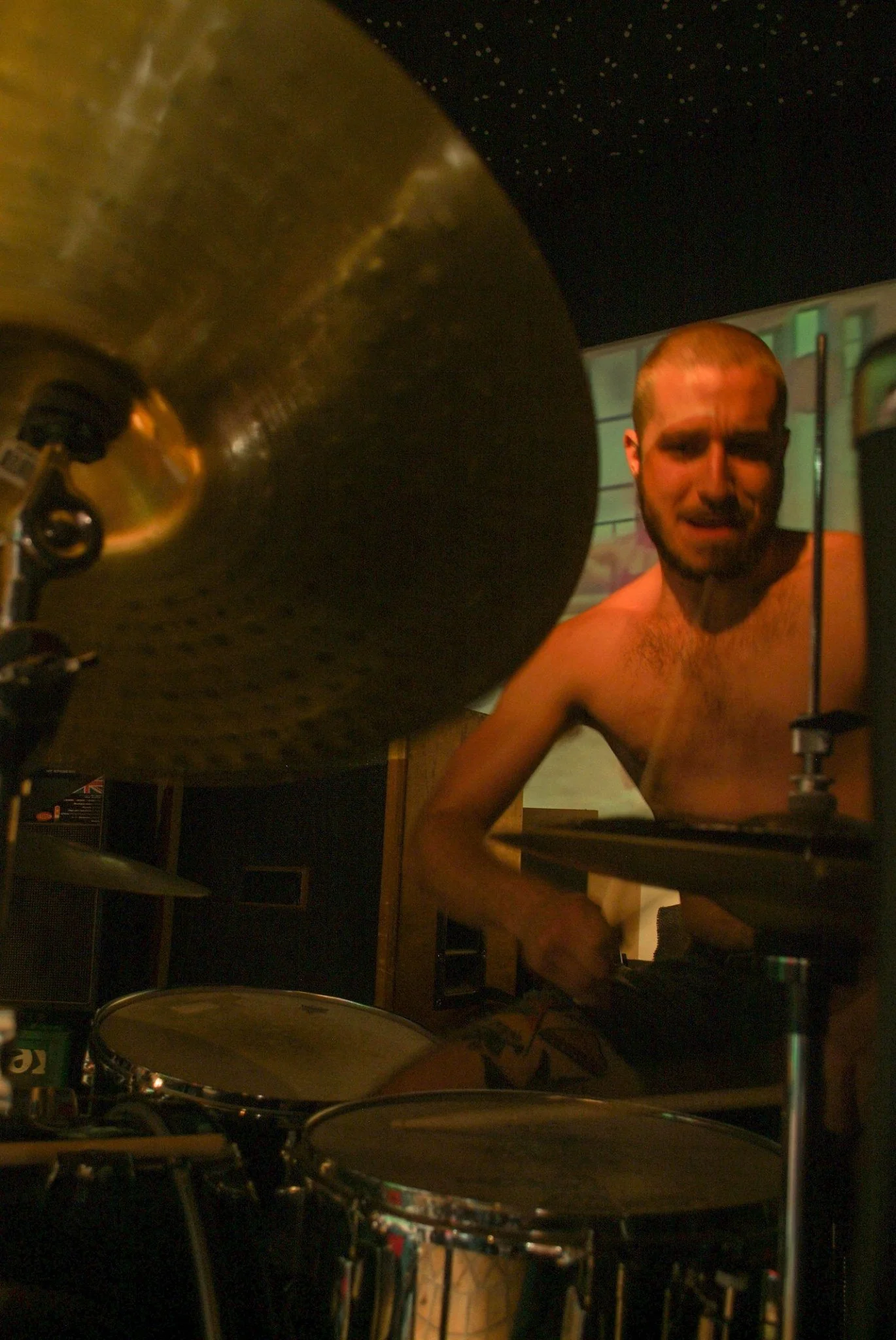 A shirtless man with a beard and short hair playing drums, with a cymbal in the foreground and a microphone stand nearby, in a dimly lit room with colorful walls.