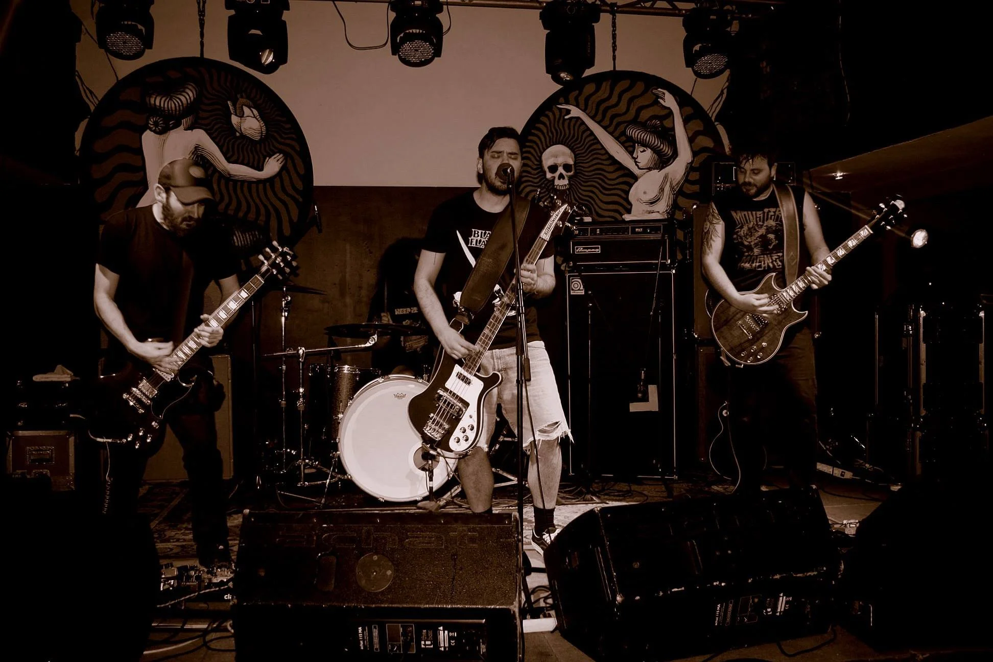 A band performs on stage with two guitarists, a drummer, and a vocalist. The stage has large circular artwork featuring stylized women, with a skull between them. The scene is dark with stage lights overhead.