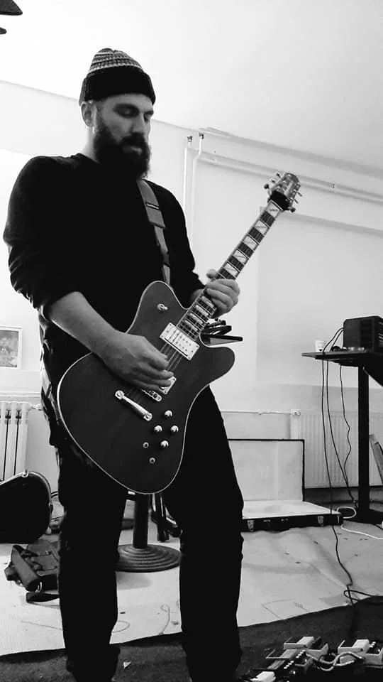 A man with a beard, wearing a beanie, playing an electric guitar in a room.