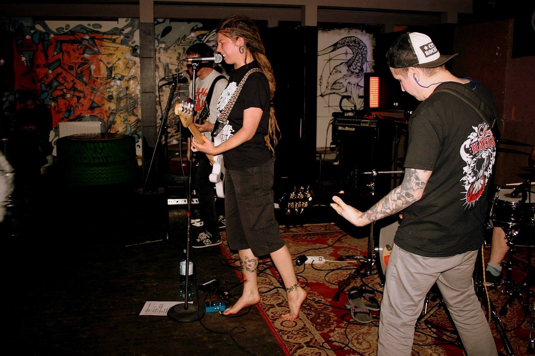Musicians performing in a lively bar or club: a female guitarist singing into a microphone with a smile, barefoot, with tattoos on her legs, wearing a black t-shirt and shorts. A male guitarist with tattoos, wearing a black cap and a black t-shirt, i