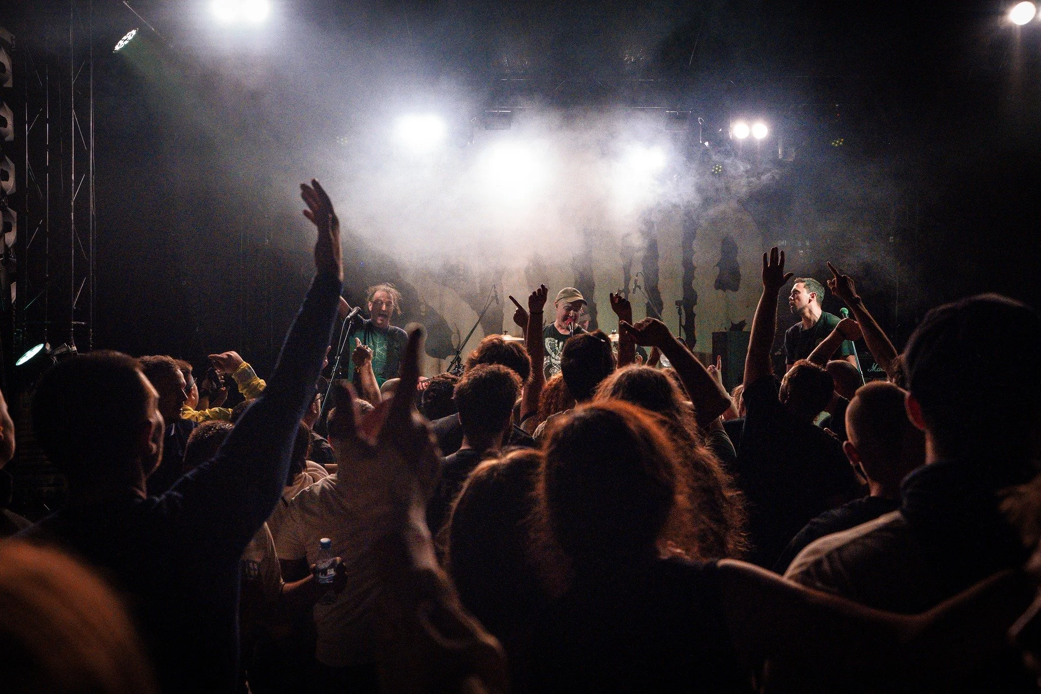 Concert scene with a crowded audience raising their hands and three performers on stage, illuminated by bright lights and fog.
