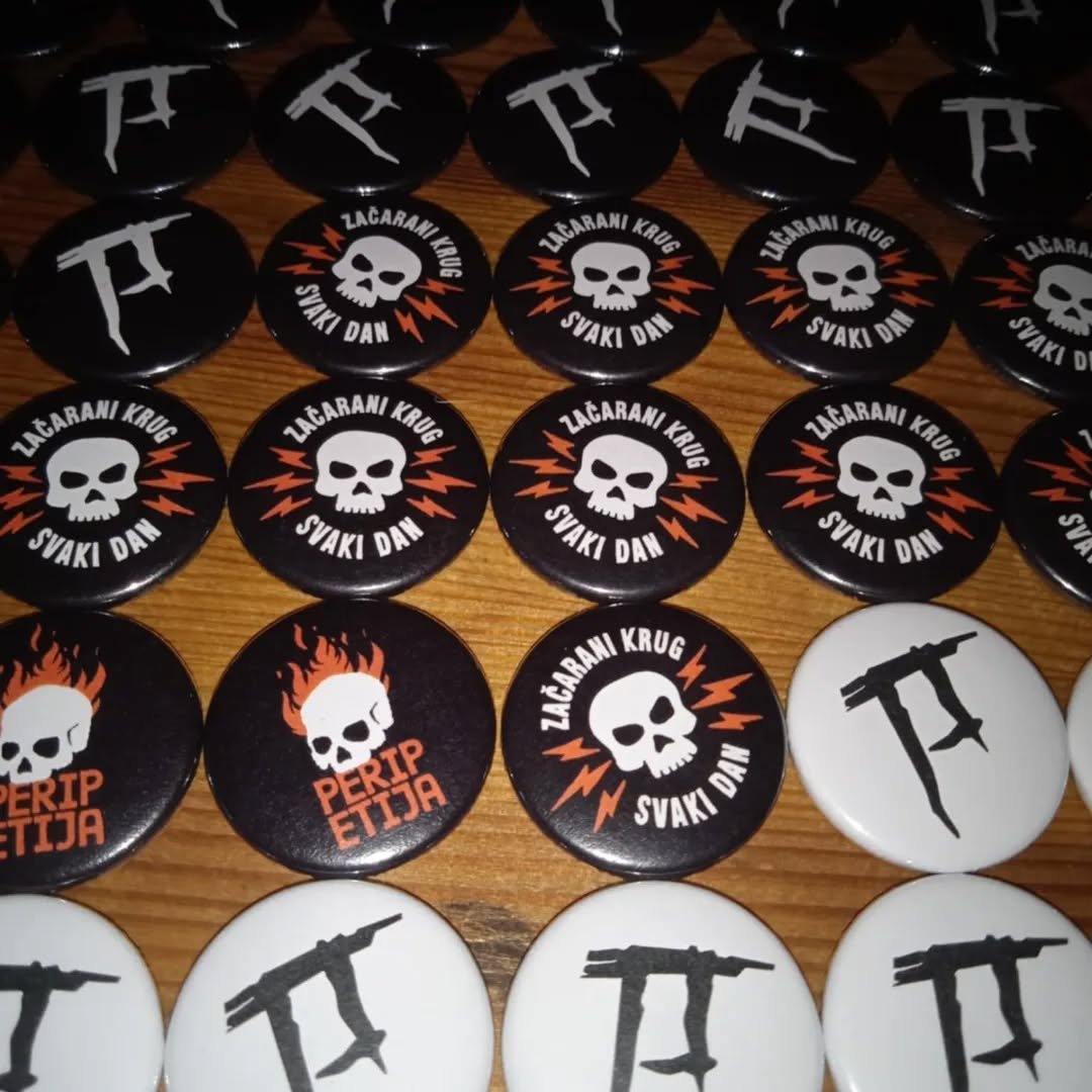 Collection of circular pins on a wooden surface, featuring skull graphics, lightning bolts, guns, and text in Croatian and English.