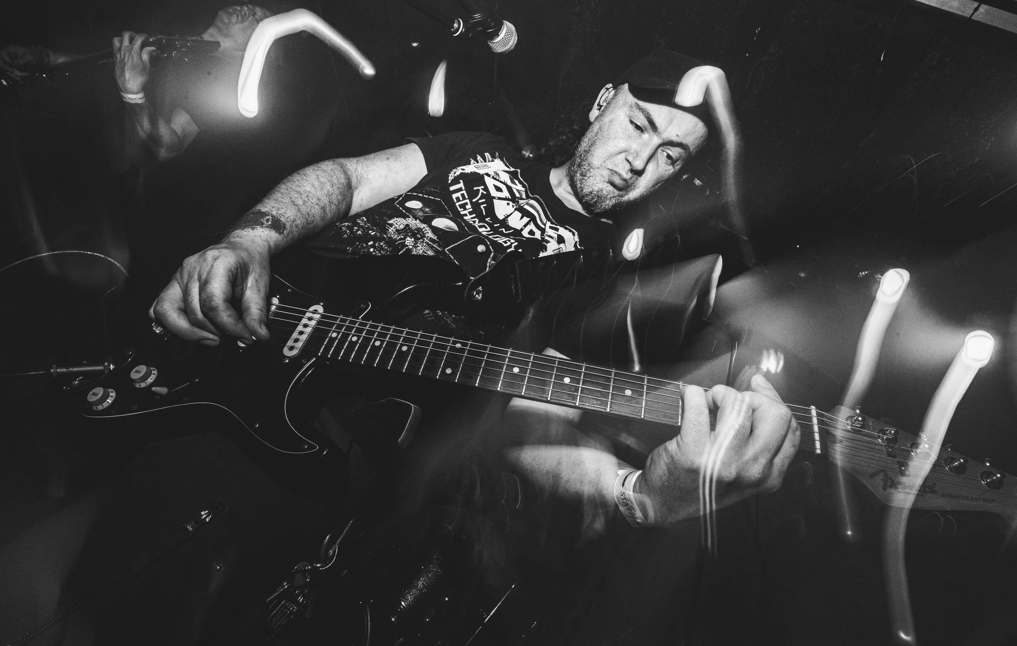 A man is lying down in a capsule-like chair, playing an electric guitar. The scene is dark with overhead lights. The man has long curly hair, wears a graphic T-shirt, and has an intense expression.