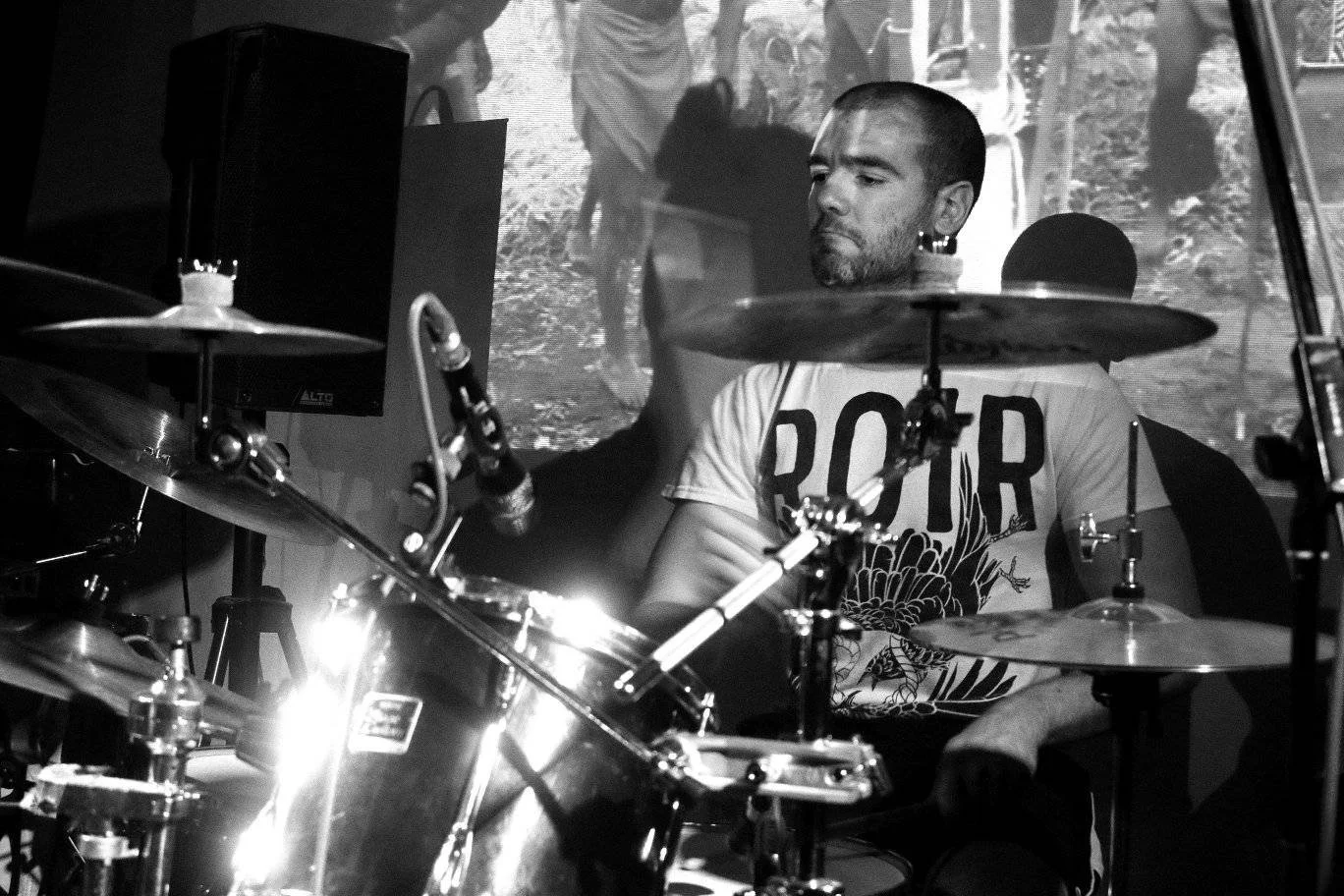 A man playing a drum set during a live performance, with a black and white background showing a scene with trees and people.