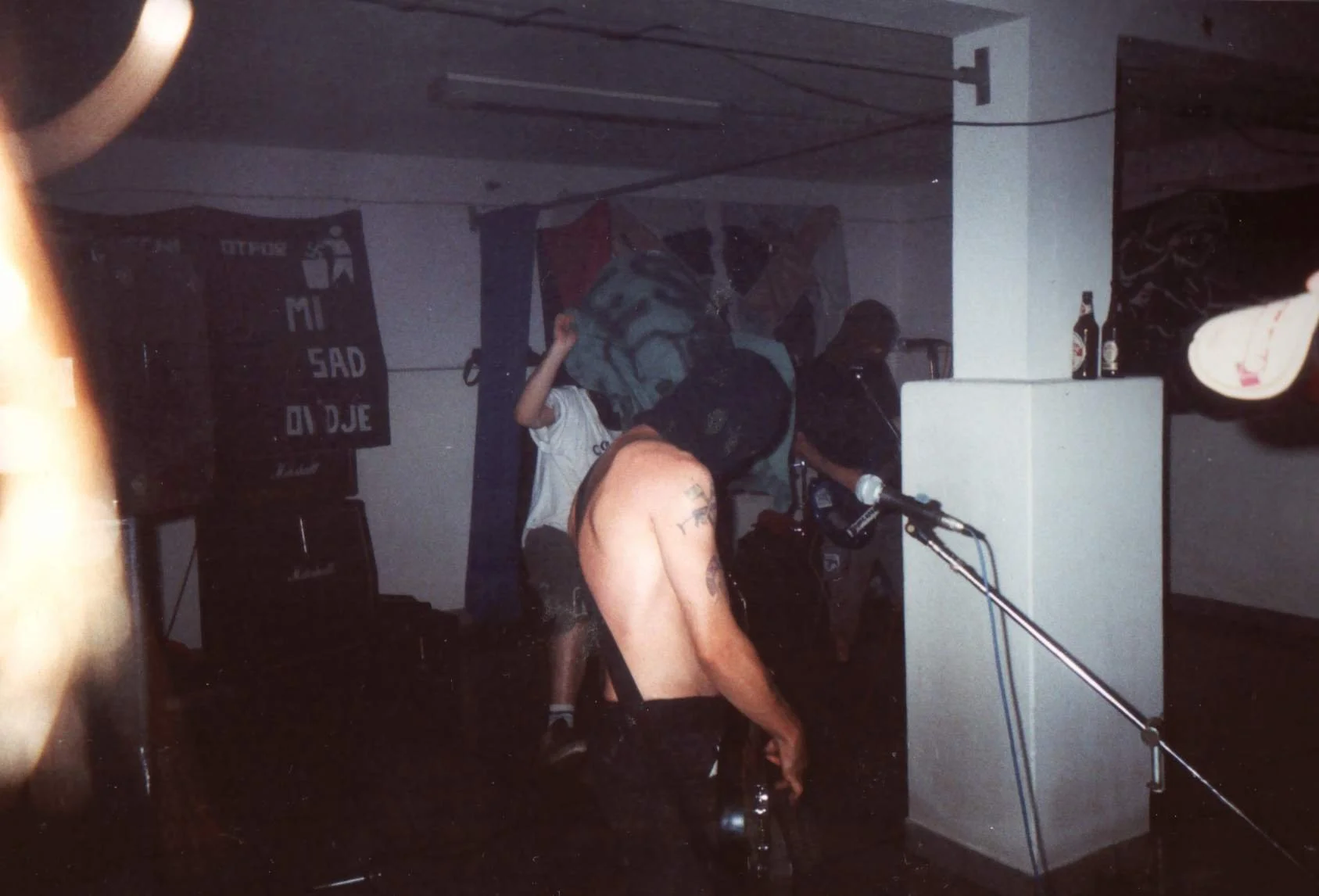 A person with tattoos on their arm, leaning over a table or stand with their shirt off, in a dimly lit room with people and equipment, possibly at a music or underground event.