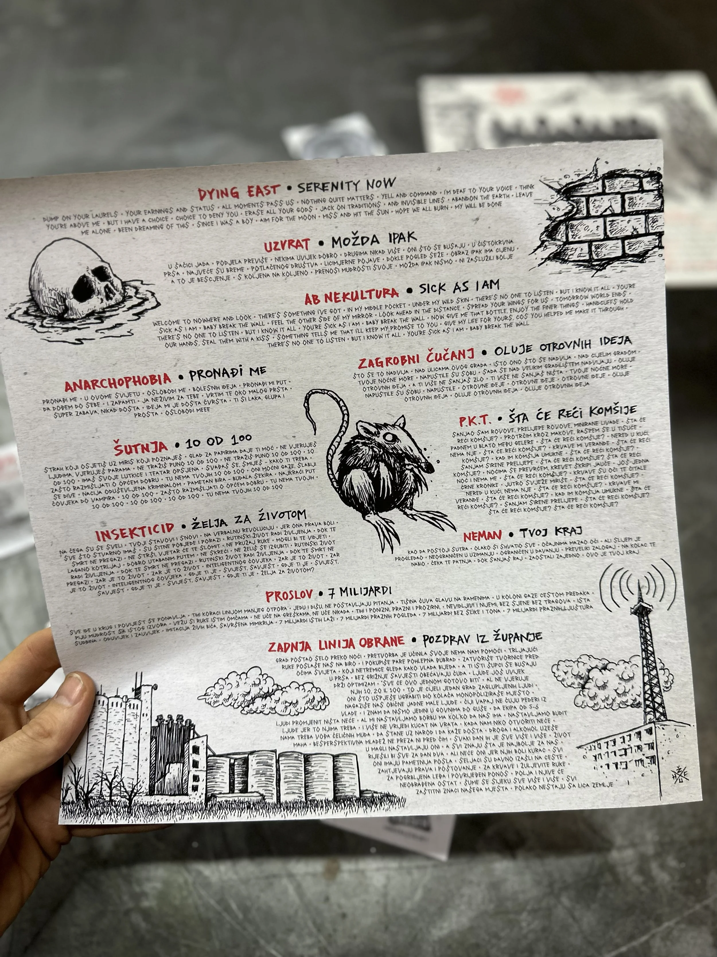 A hand holding a black and white illustrated newspaper or flyer with handwritten-style text and drawings, including a small skull, a bird, and a cityscape. The paper features various titles and sections written in a language using Latin script.