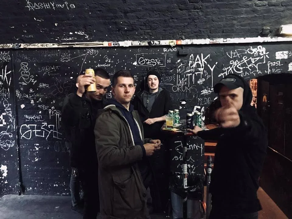 Four young men standing in a graffiti-covered nightclub or bar, with drinks on a small table in the background, some making gestures and looking at the camera.