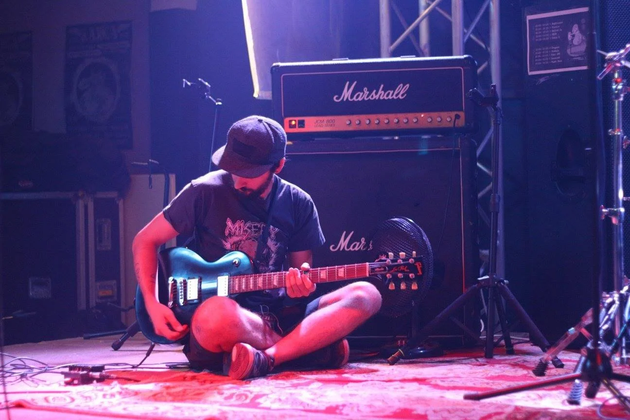 A person sitting on the floor playing an electric guitar in a music stage setting with Marshall amplifiers behind him.