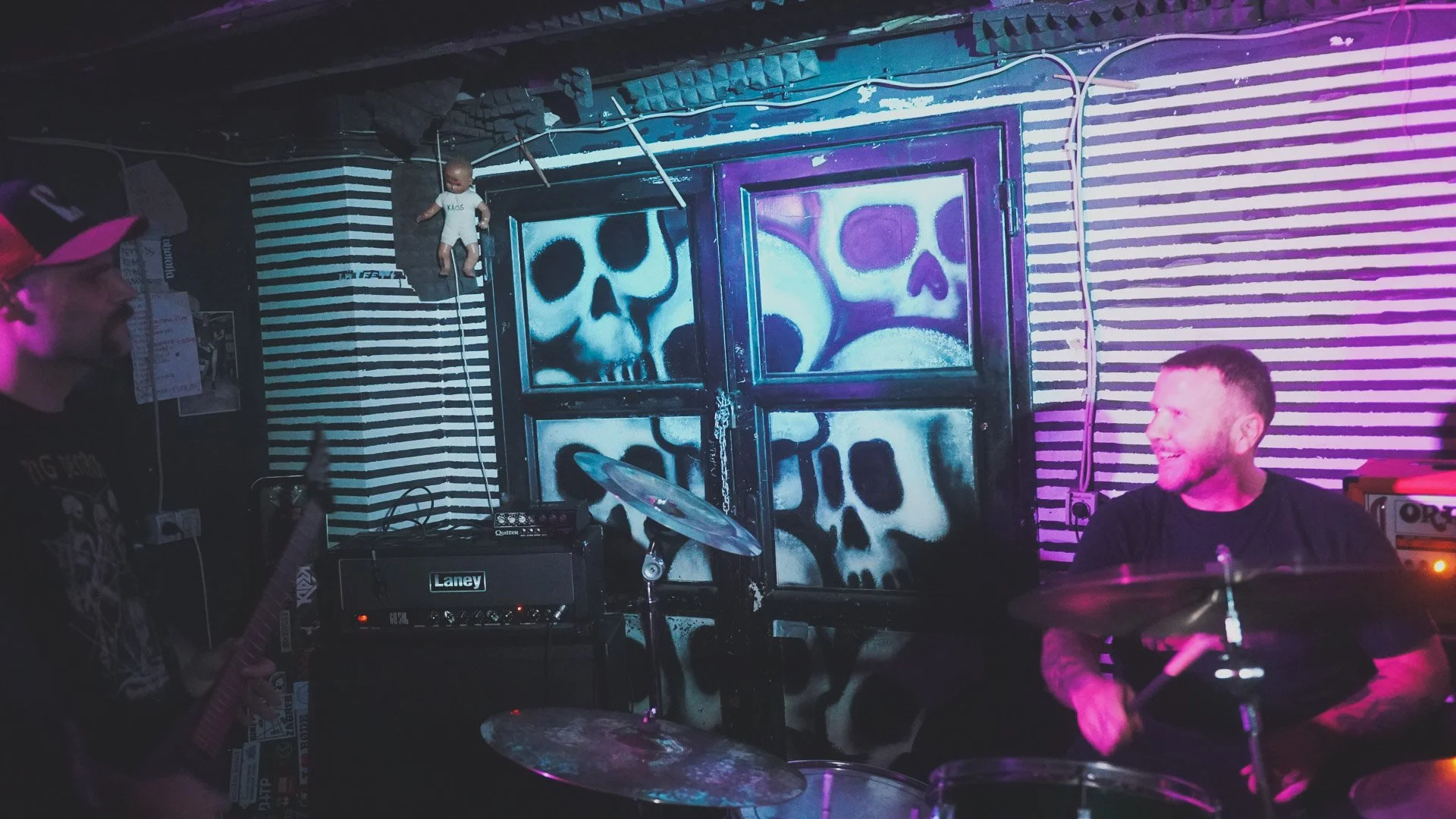 People performing music in a dark room with skull artwork on the back wall, colorful lighting, musical equipment, and a person playing drums.