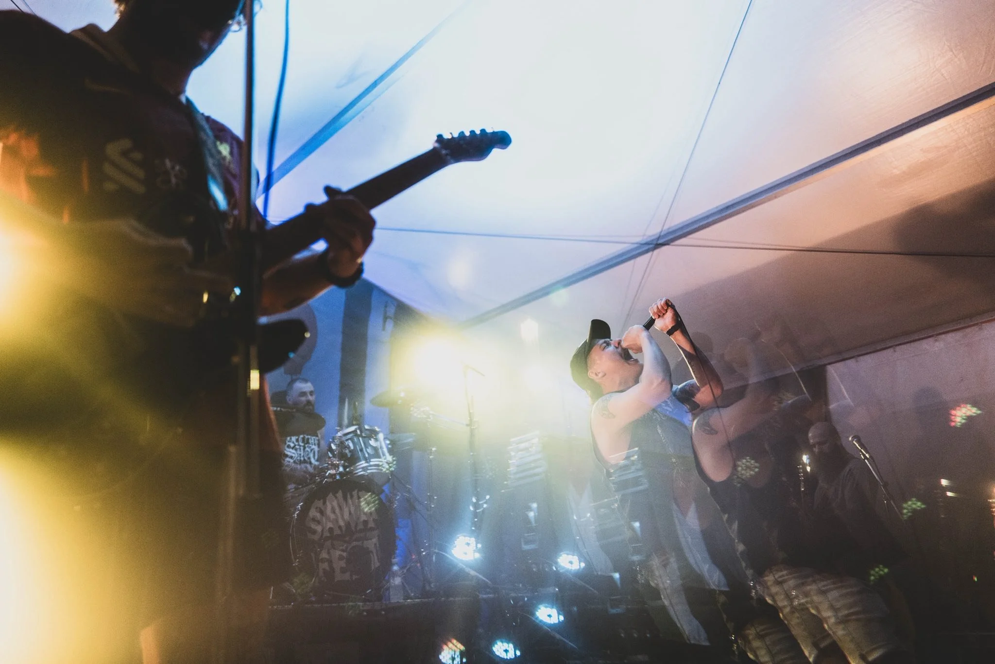 A musician with tattoos on his arm and wearing a cap, singing into a microphone during a live performance, with a band and colorful stage lighting in the background.