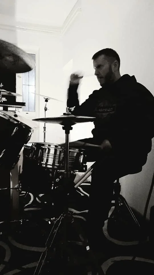 A man with a beard playing a drum set indoors, captured in black and white.