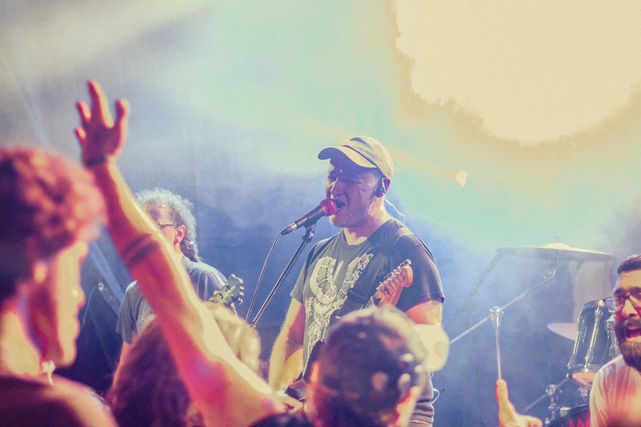 Musicians perform on stage at a concert, with a lead singer playing guitar and singing into a microphone, surrounded by audience members raising their hands.