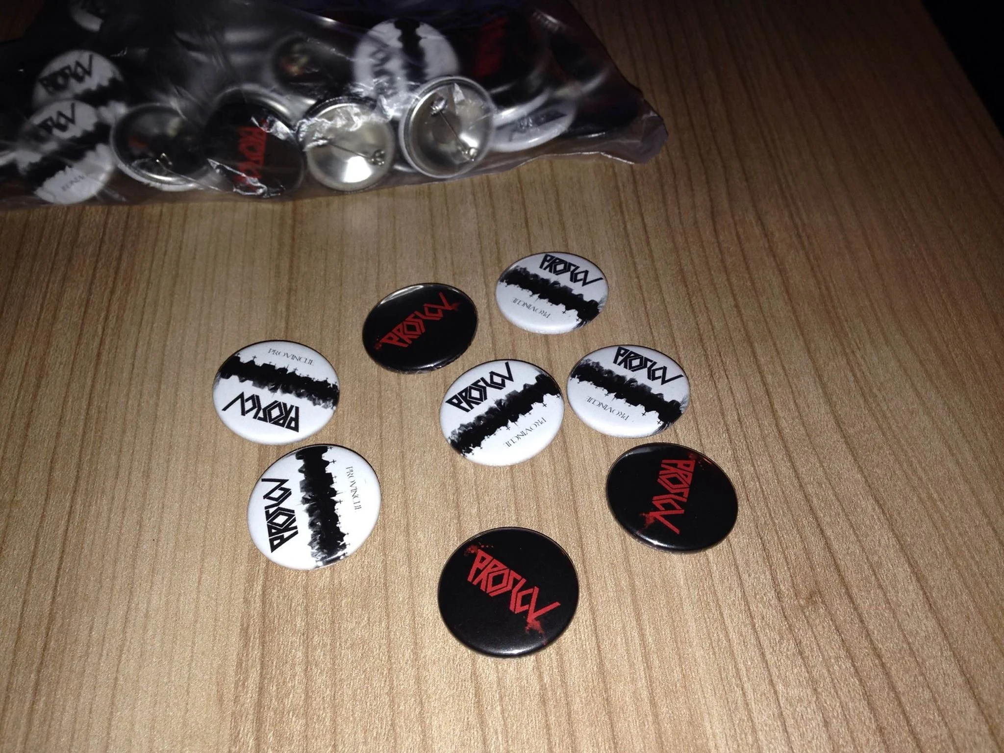 A wooden table with several round buttons featuring the word "PROSLOV" in black and red text, some buttons are inside a plastic bag with similar buttons visible.