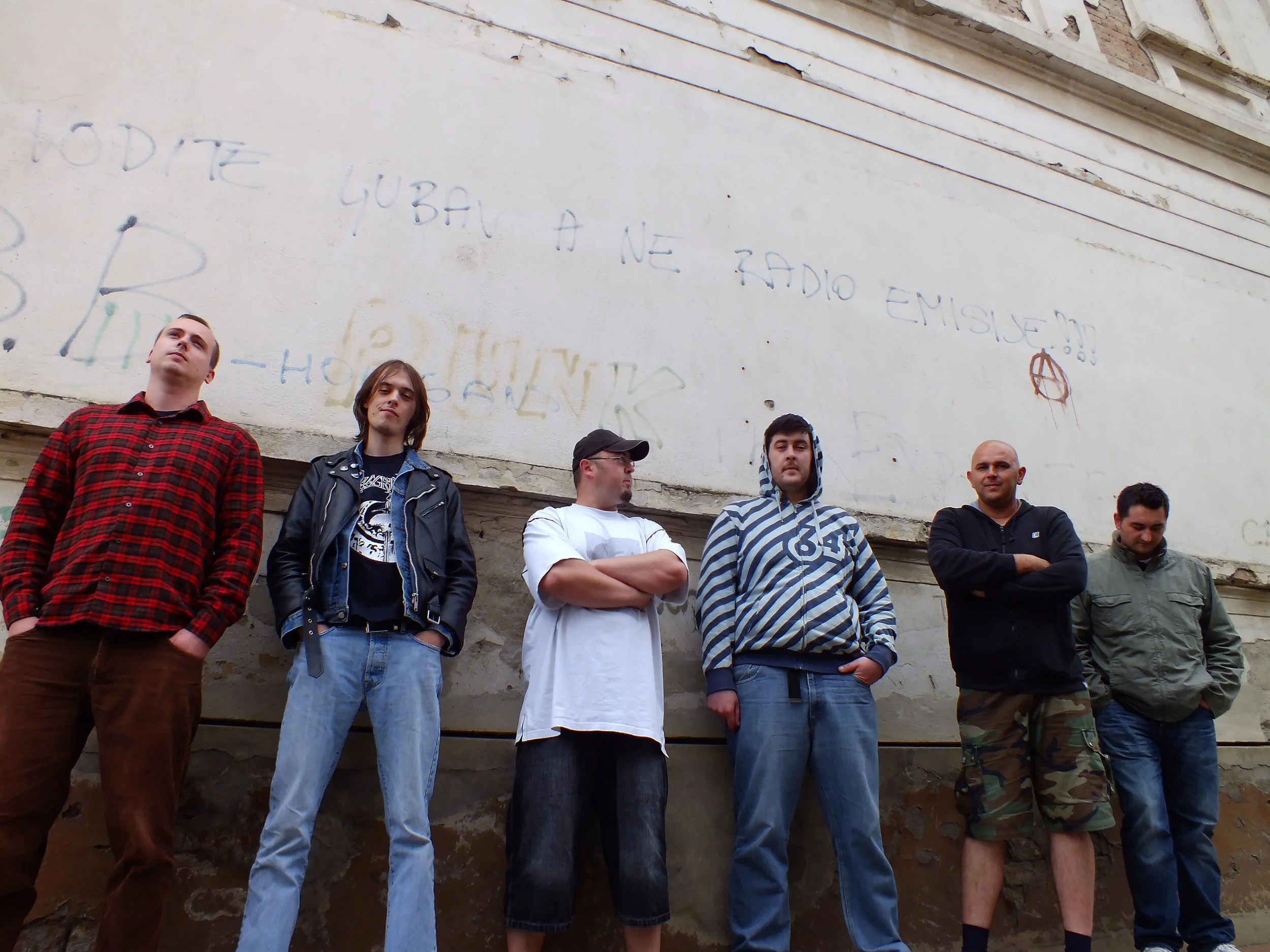 A group of seven young men standing in front of a weathered, graffiti-covered wall.