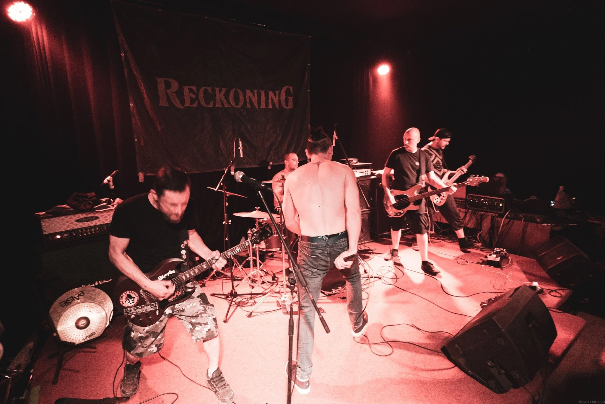 A band of musicians preparing for a performance on stage with a large banner that reads 'RECKONING' in the background. The stage is lit with red lighting, and there are multiple music instruments and equipment present, including guitars, drums, and a