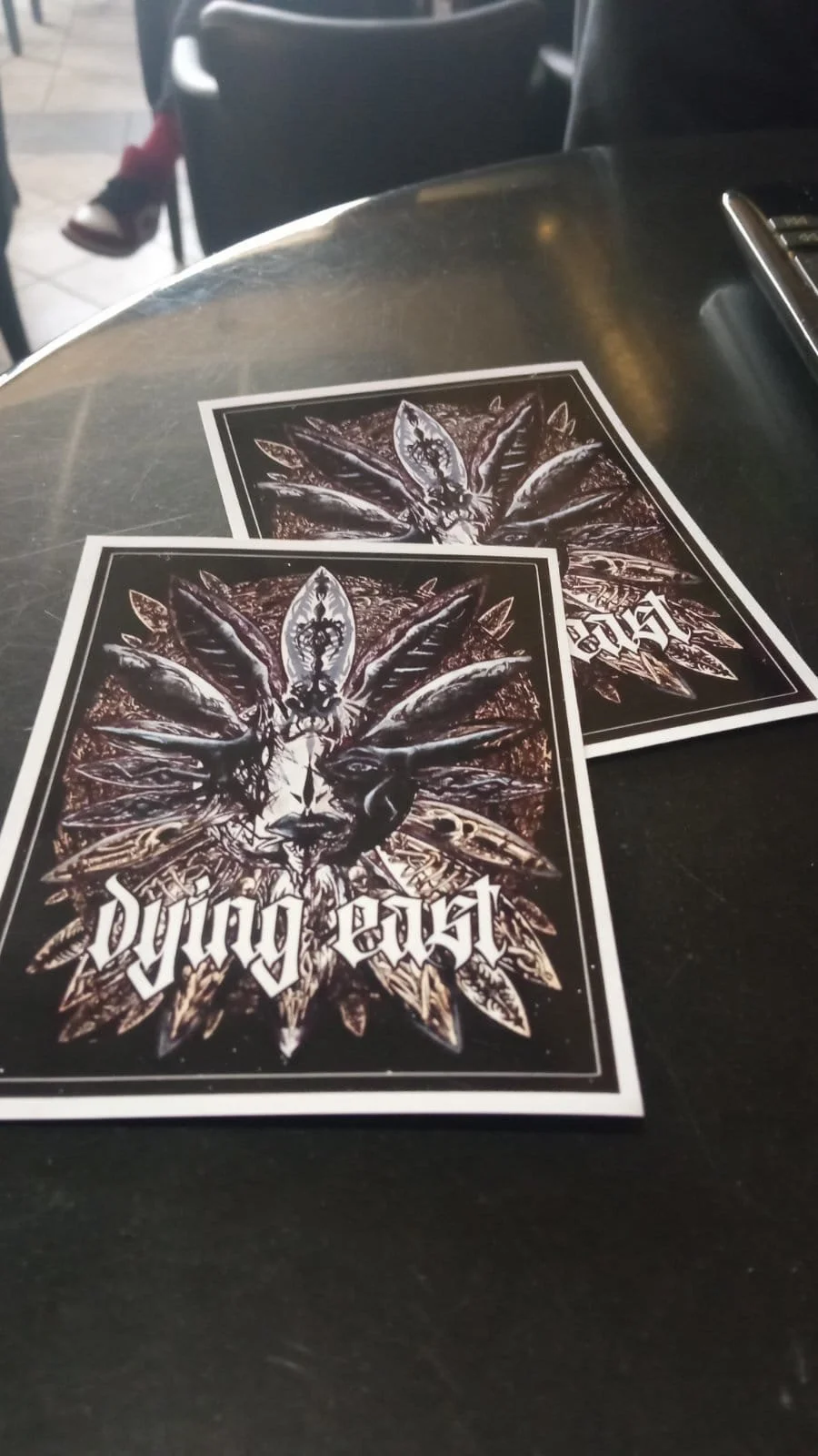Two promotional flyers on a dark table featuring a graphic of a goat with multiple horns and wings, with the words "dying beast" in a gothic font.