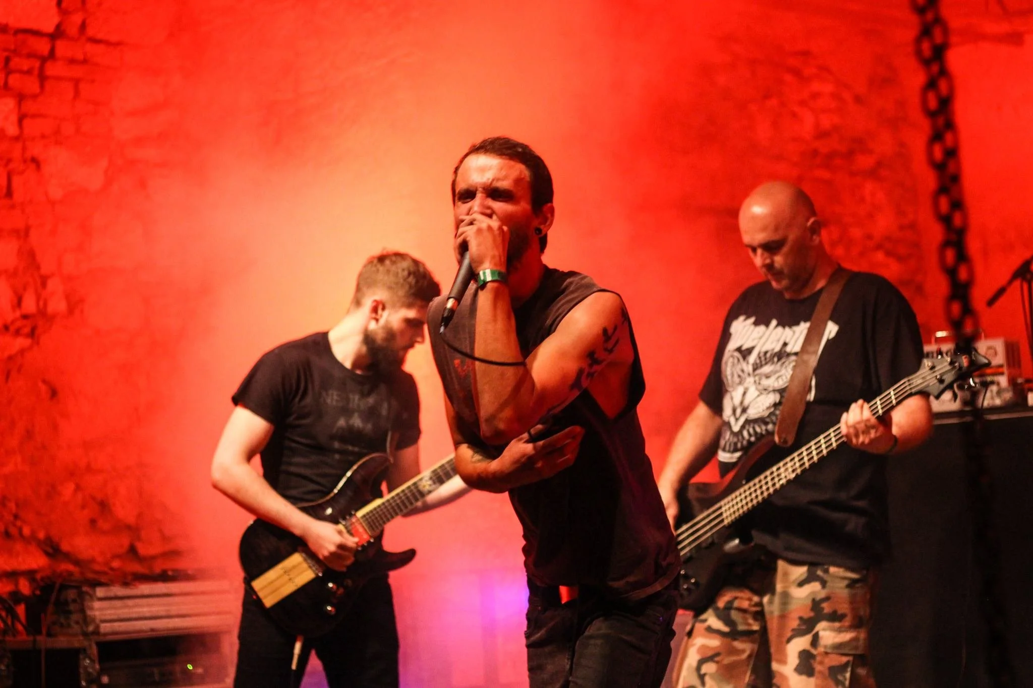 A band performing on stage with a lead singer holding a microphone, two guitarists playing electric guitars, and a drummer in the background, all against a red wall.