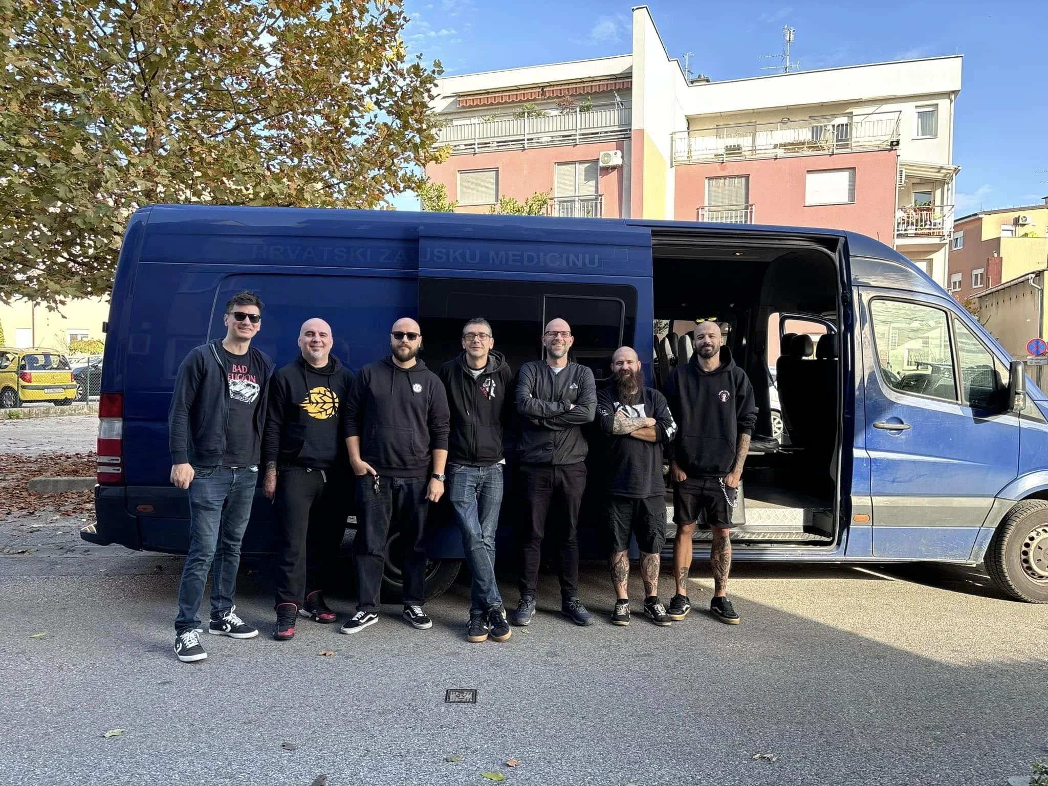 A group of seven men standing in front of a blue van on a parking lot with residential buildings and trees in the background.